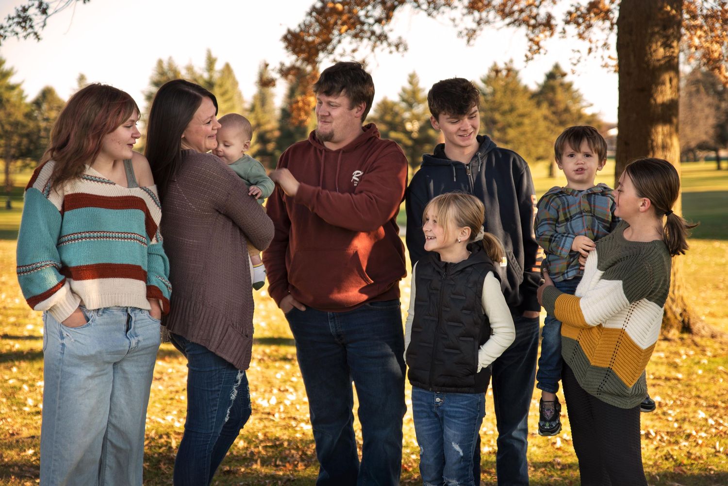 A joyful family of eight poses together in a park with warm golden autumn light.