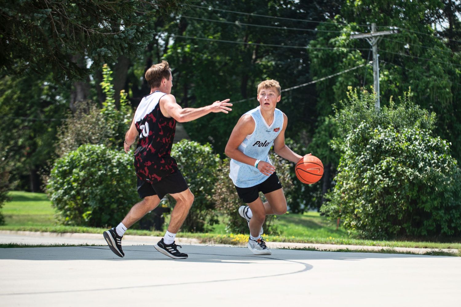 Two young men playing outdoor basketball on a sunny day, one dribbling past a defender on a paved court.