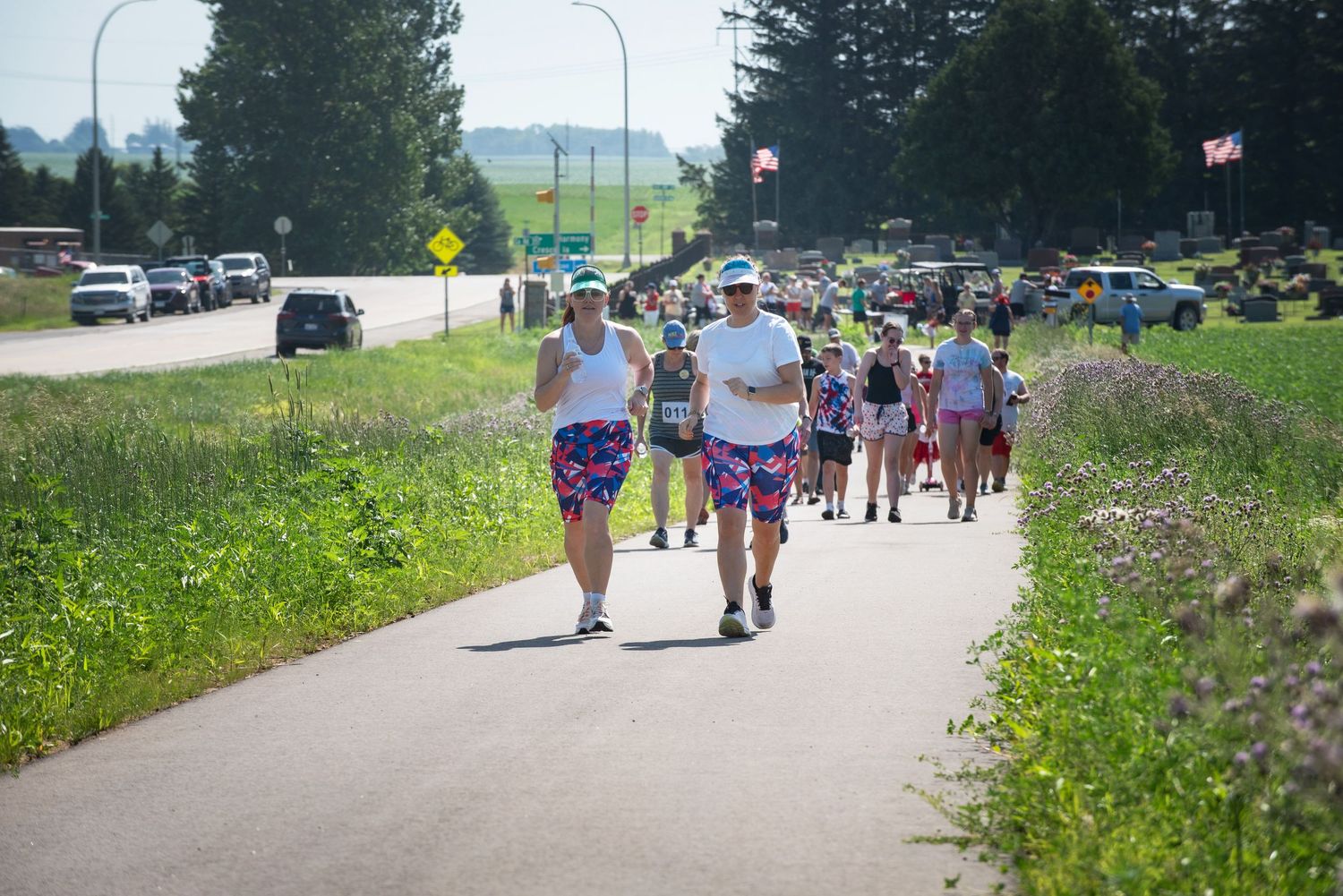 People in colorful patterned shorts walking and running on paved path on sunny summer day at Harmony MN 4th of July Celebration..
