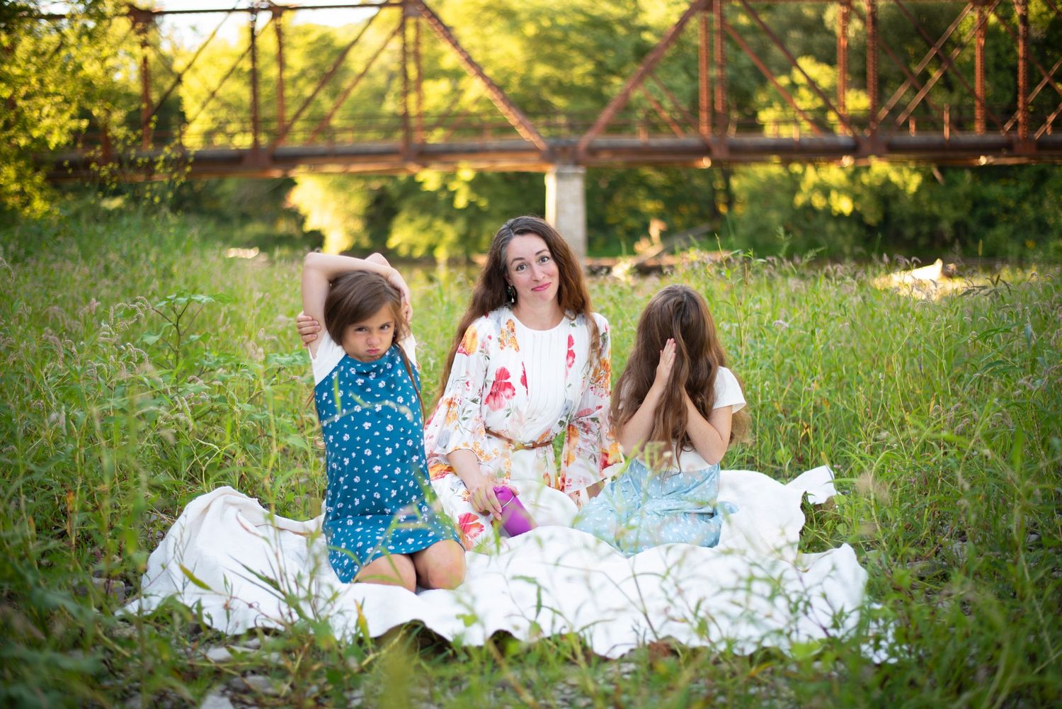 A mother and two daughters sitting together on a white blanket in a lush green meadow.