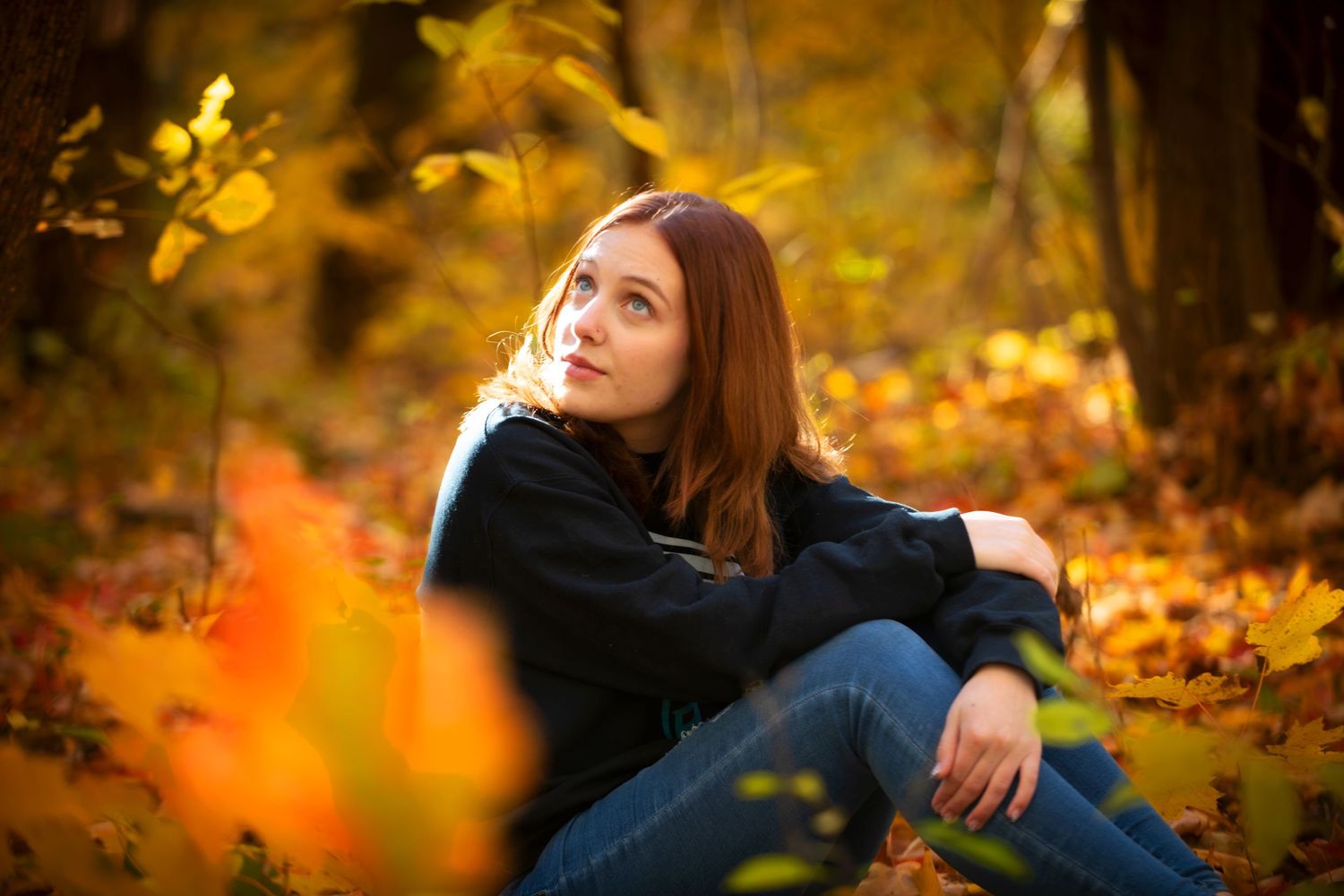 Young woman sitting among golden autumn leaves in a forest, gazing upward thoughtfully.