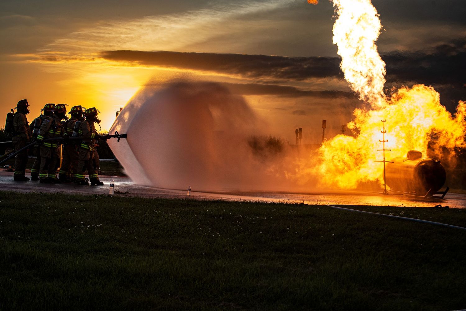 Firefighters spraying water on a large practice fire at dramatic sunset.
