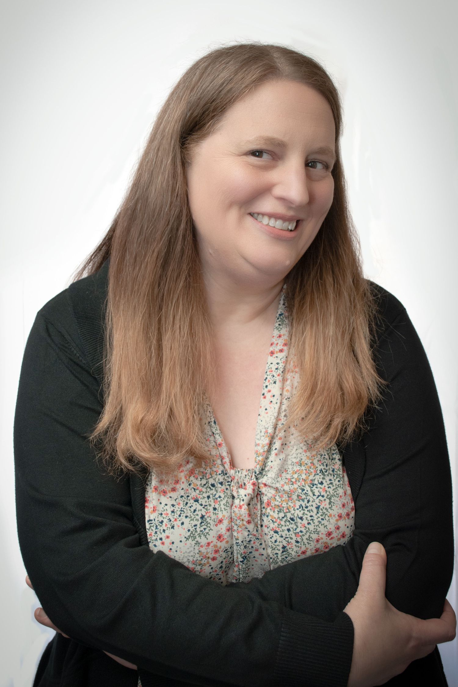Smiling woman with long auburn hair wearing a floral blouse and black cardigan poses with arms crossed.