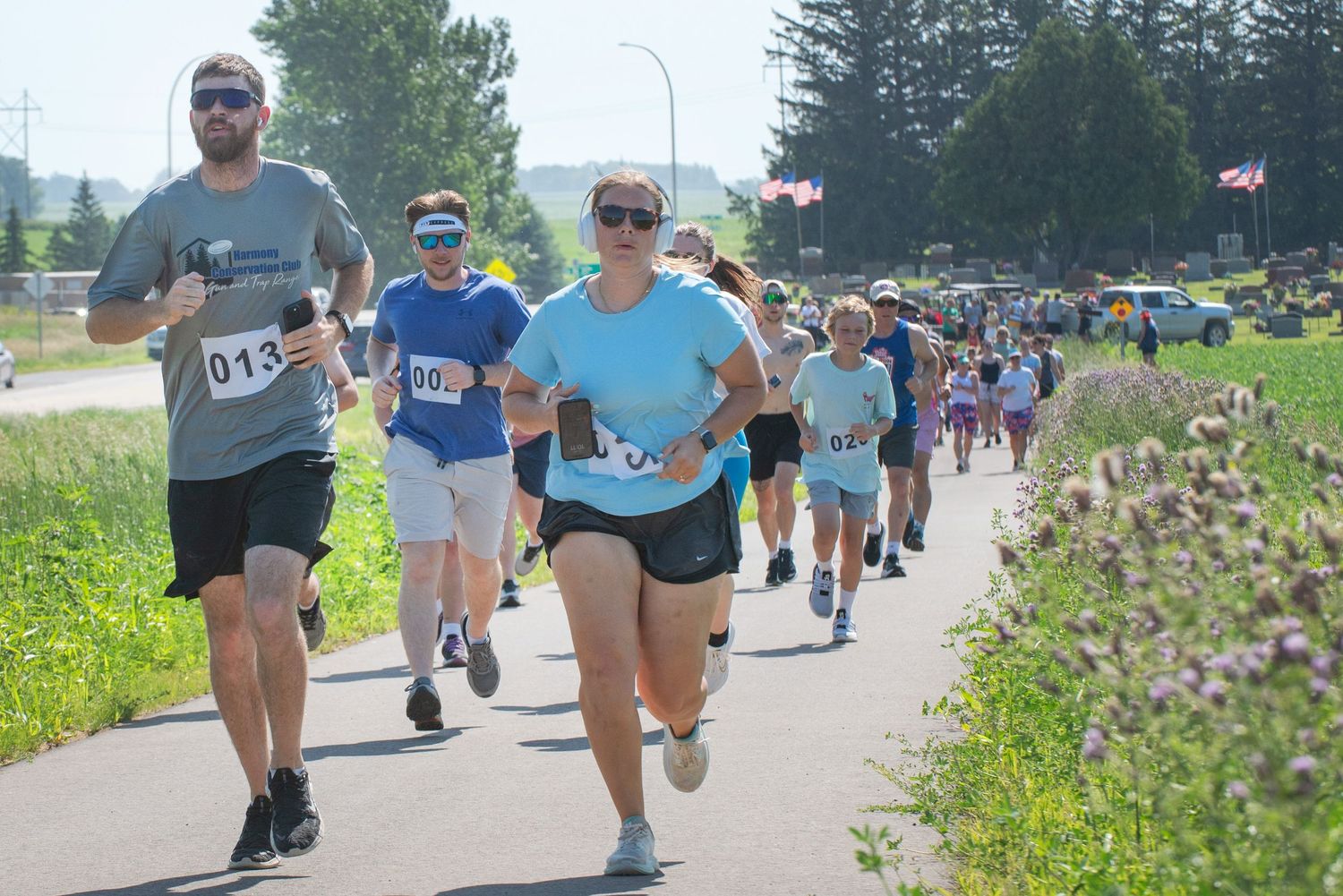 Group of runners with race bibs running on paved path on bright sunny summer day at Harmony MN 4th of July Celebration..