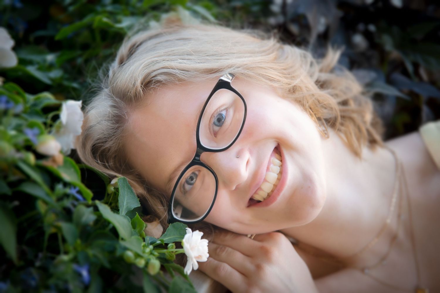 Smiling blonde girl wearing black glasses surrounded by green foliage and white flowers.
