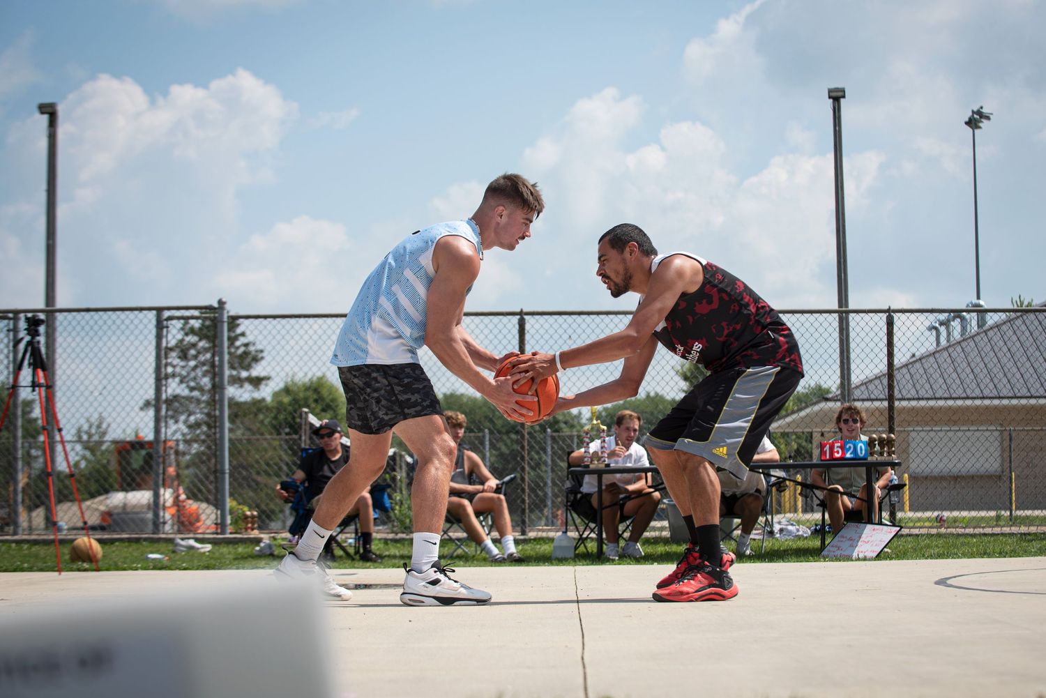 Two players battle for basketball possession on outdoor fenced court on sunny day.
