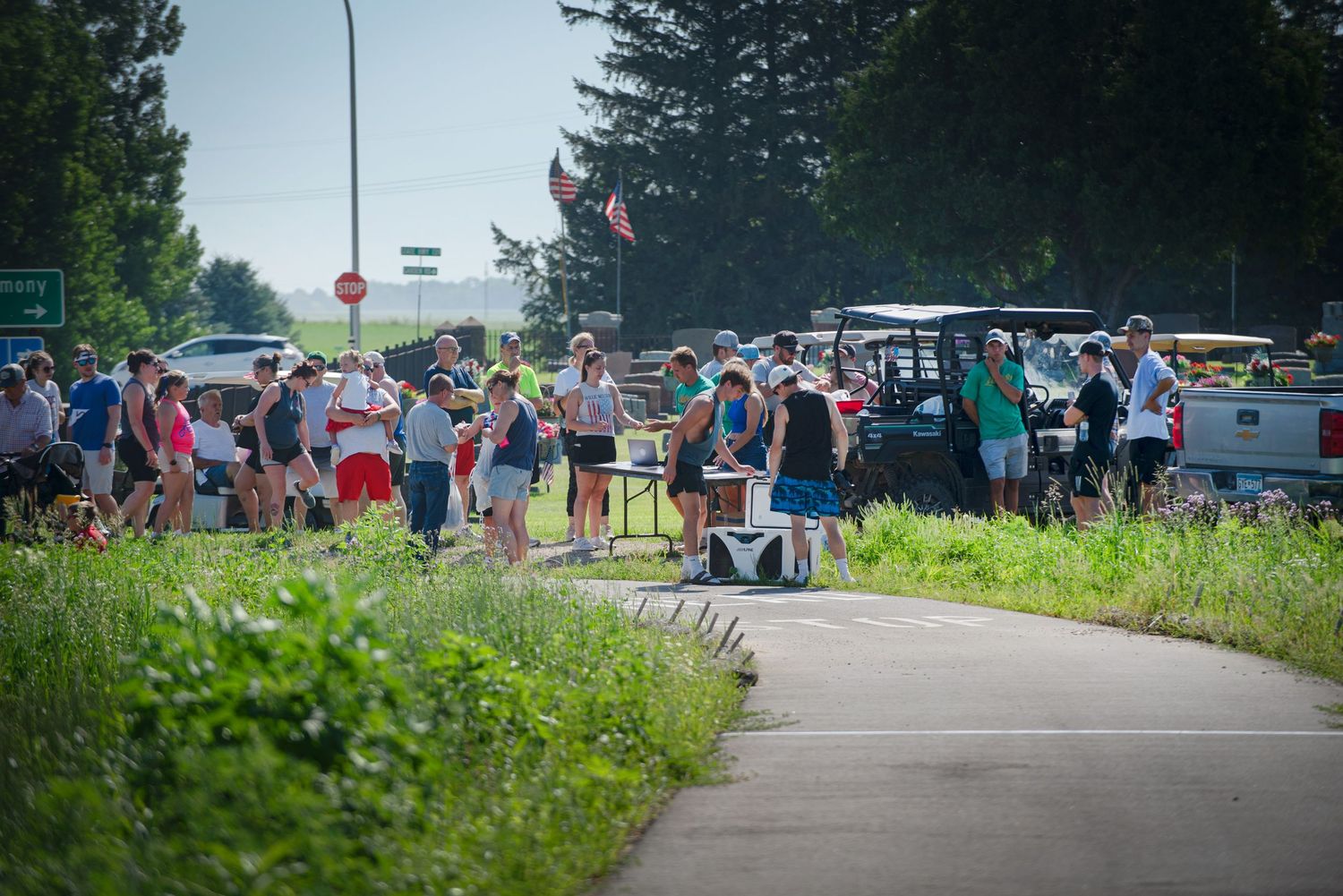 Crowd of people gathered on pathway near road on sunny summer day with American flags visible at Harmony MN 4th of July Celebration..