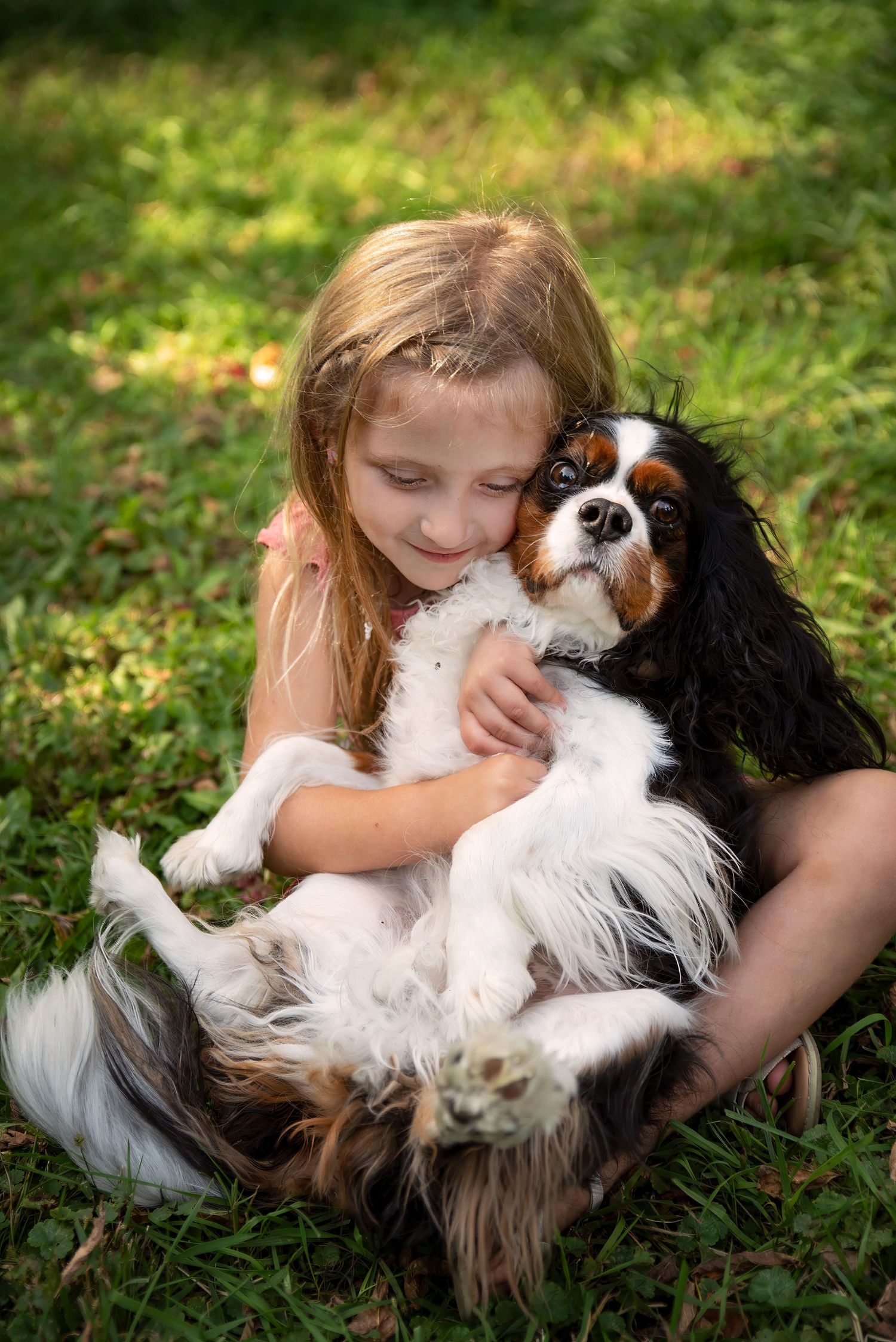 A young blonde girl lovingly hugs a Cavalier King Charles Spaniel on green grass.