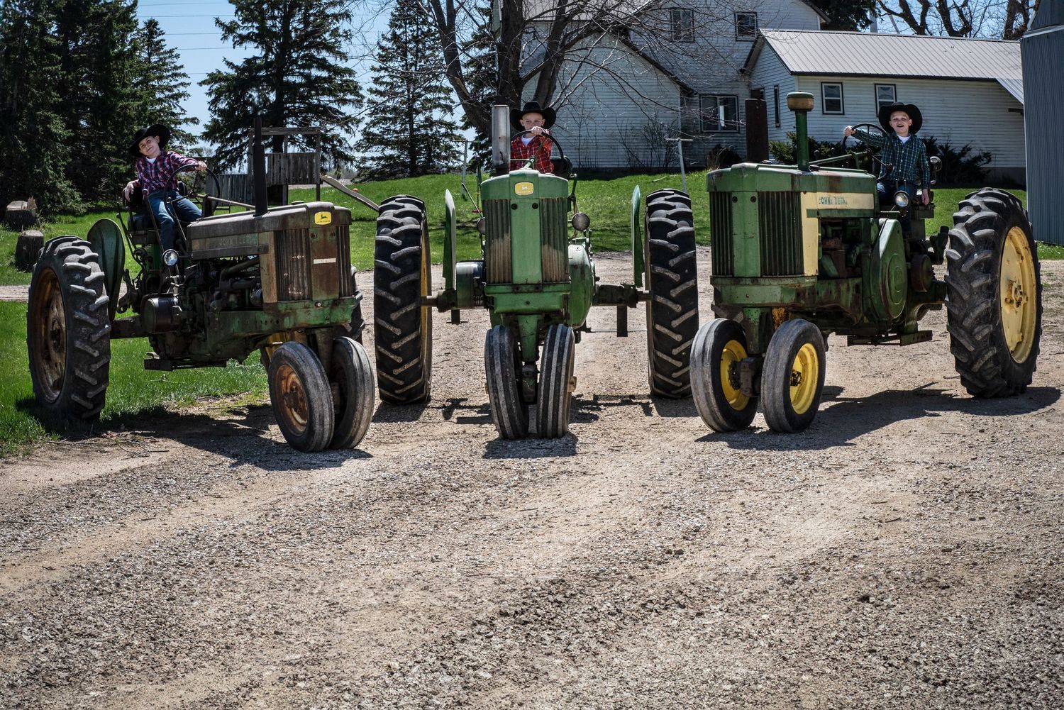Three people sitting on vintage green John Deere tractors lined up on a gravel farm driveway.