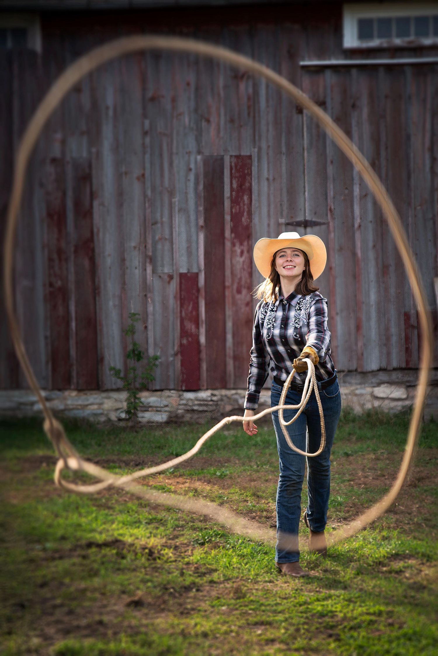 Smiling cowgirl in plaid shirt and straw hat twirling a lasso in front of a rustic barn.