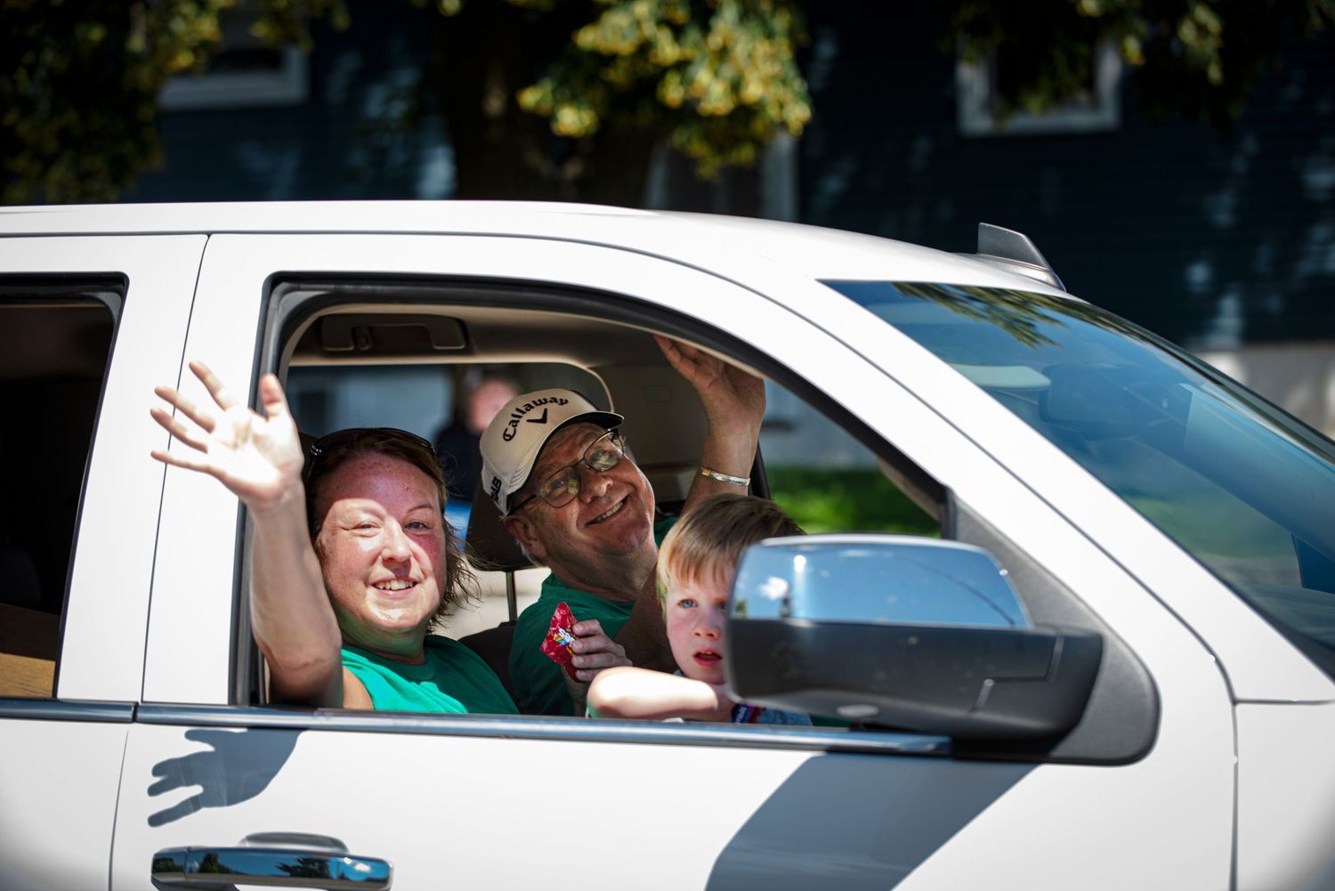 Smiling people wave from a white pickup truck window while participating in a sunny Fourth of July parade Harmony Mn.