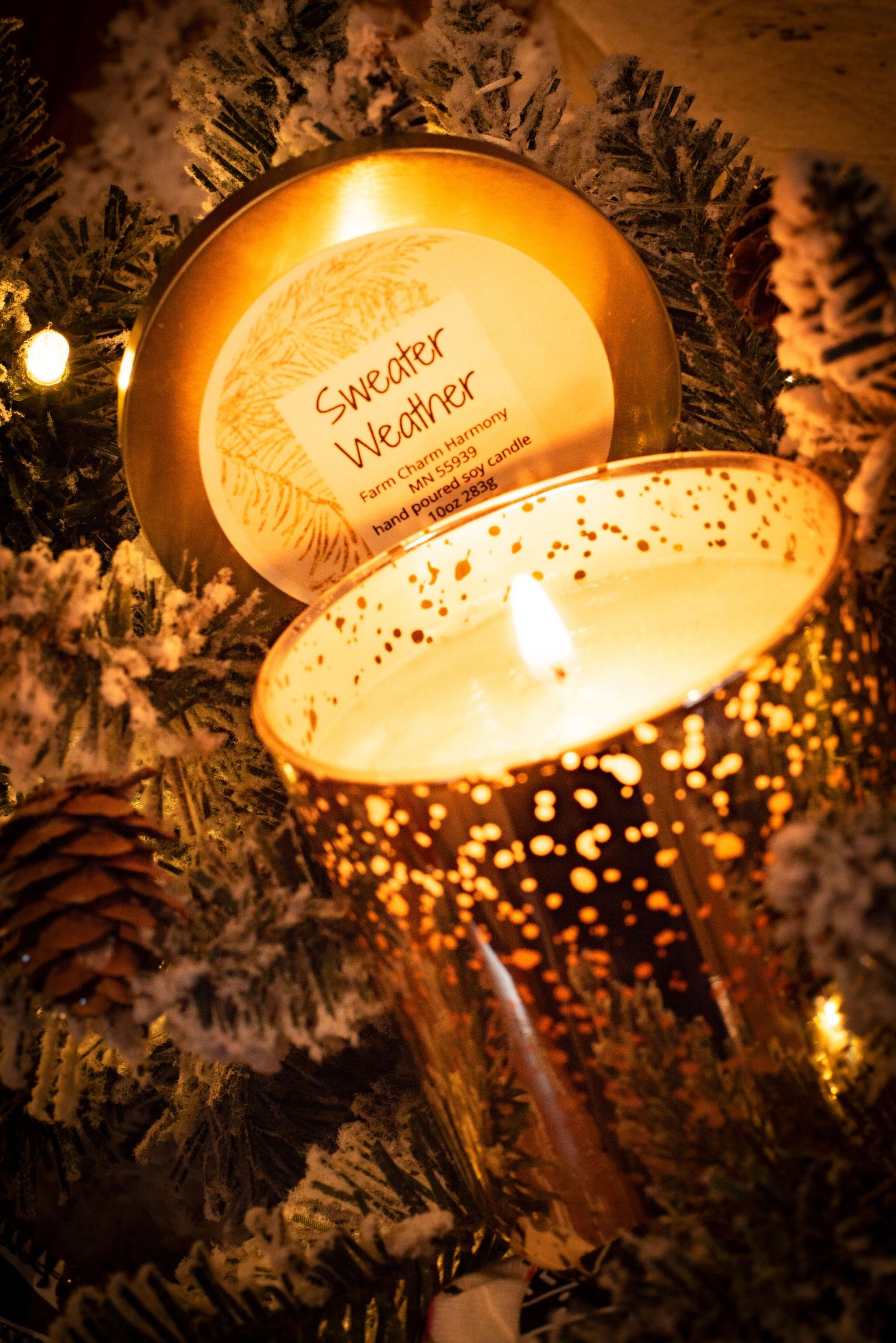 Glowing Sweater Weather scented candle nestled among snow-dusted Christmas tree branches with warm bokeh lights from Farm Charm.