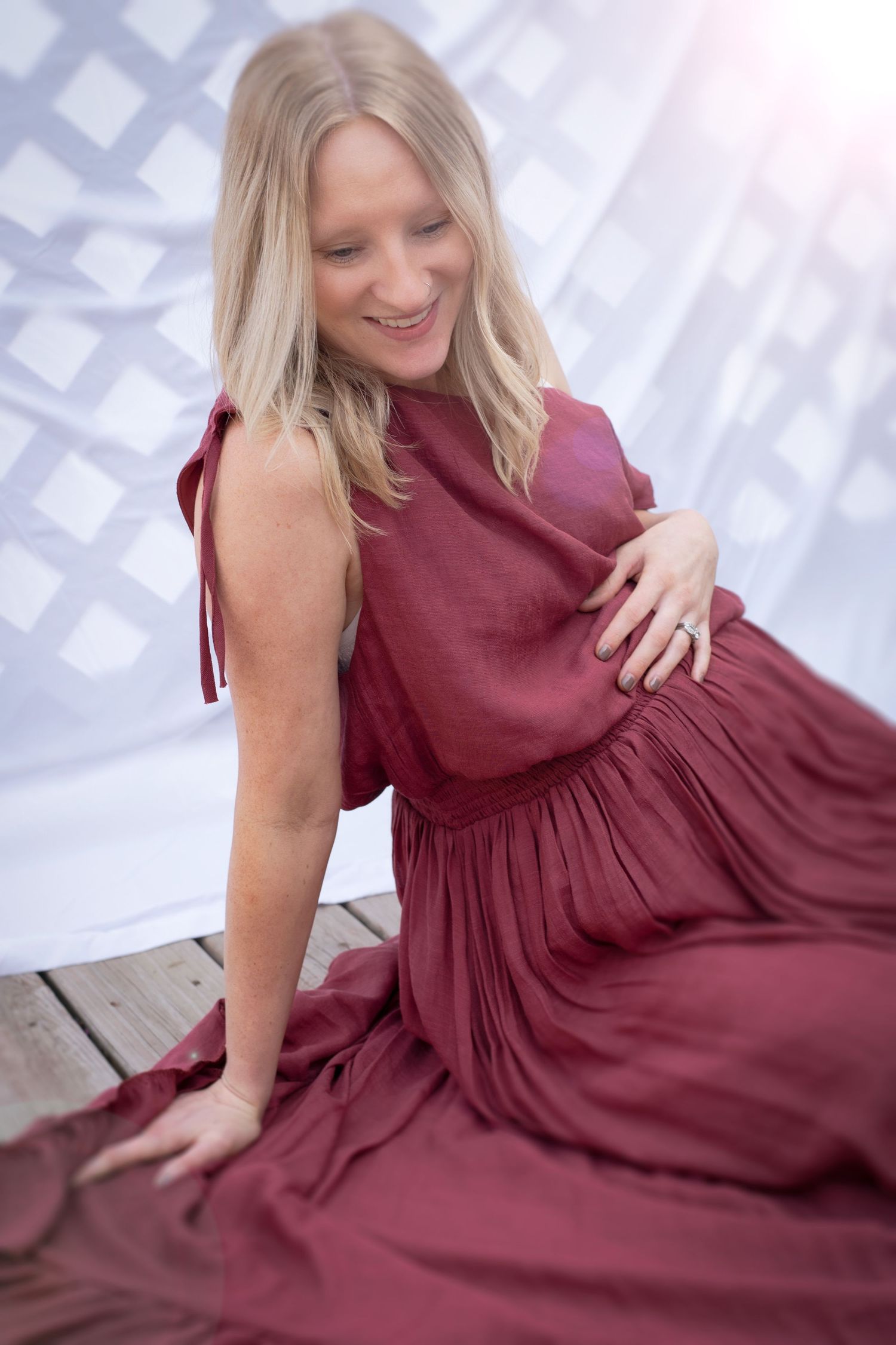 Smiling pregnant woman in burgundy dress sitting on wooden deck outdoors.
