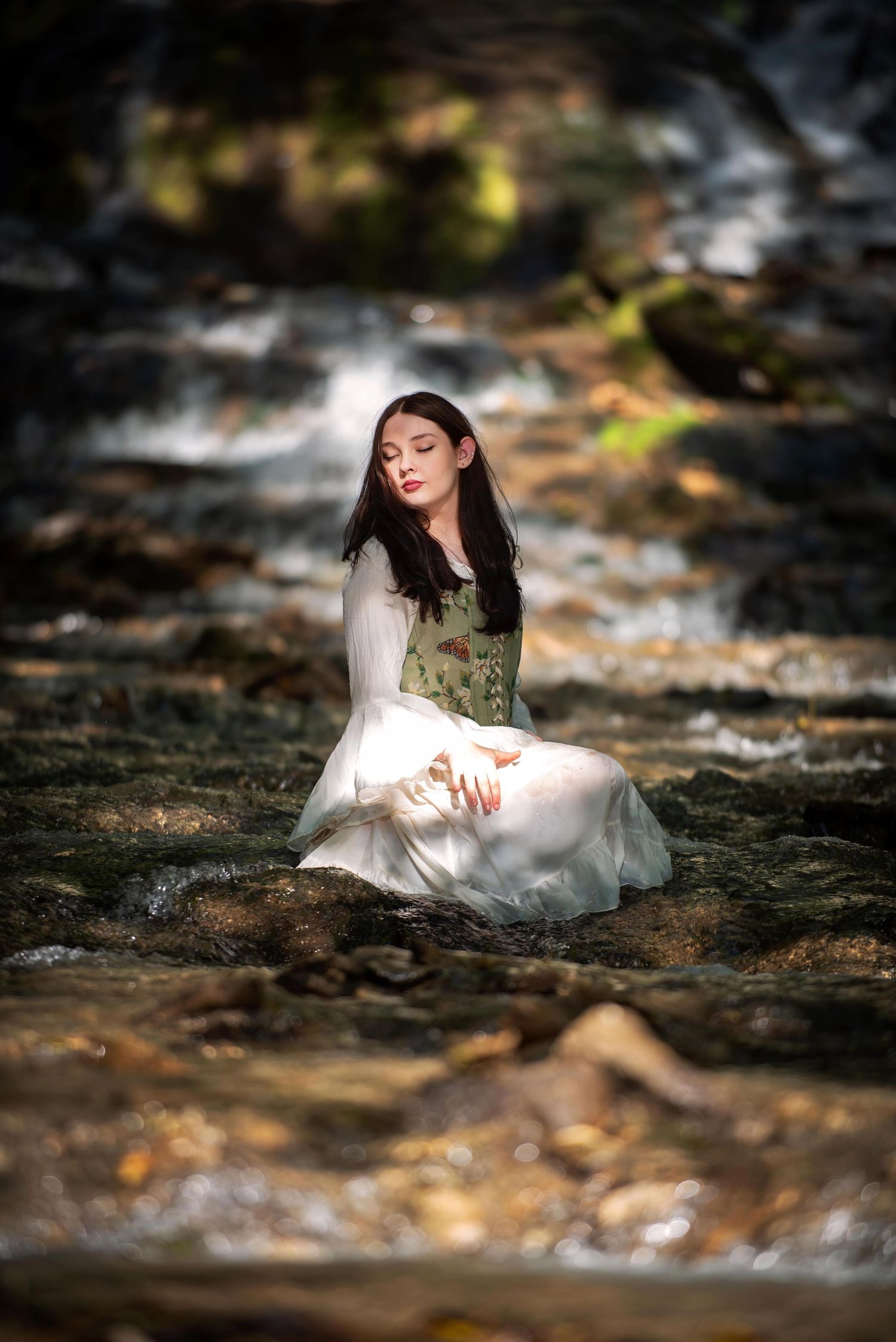 Young woman in a white medieval gown sitting peacefully on rocks beside a flowing woodland stream.