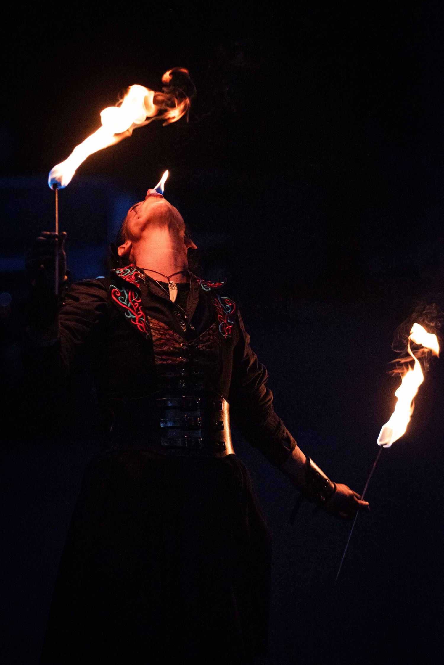 A fire eater in a black embroidered coat balances flaming torches during a dramatic nighttime performance.Outside Monster 
Bash Harmony MN.