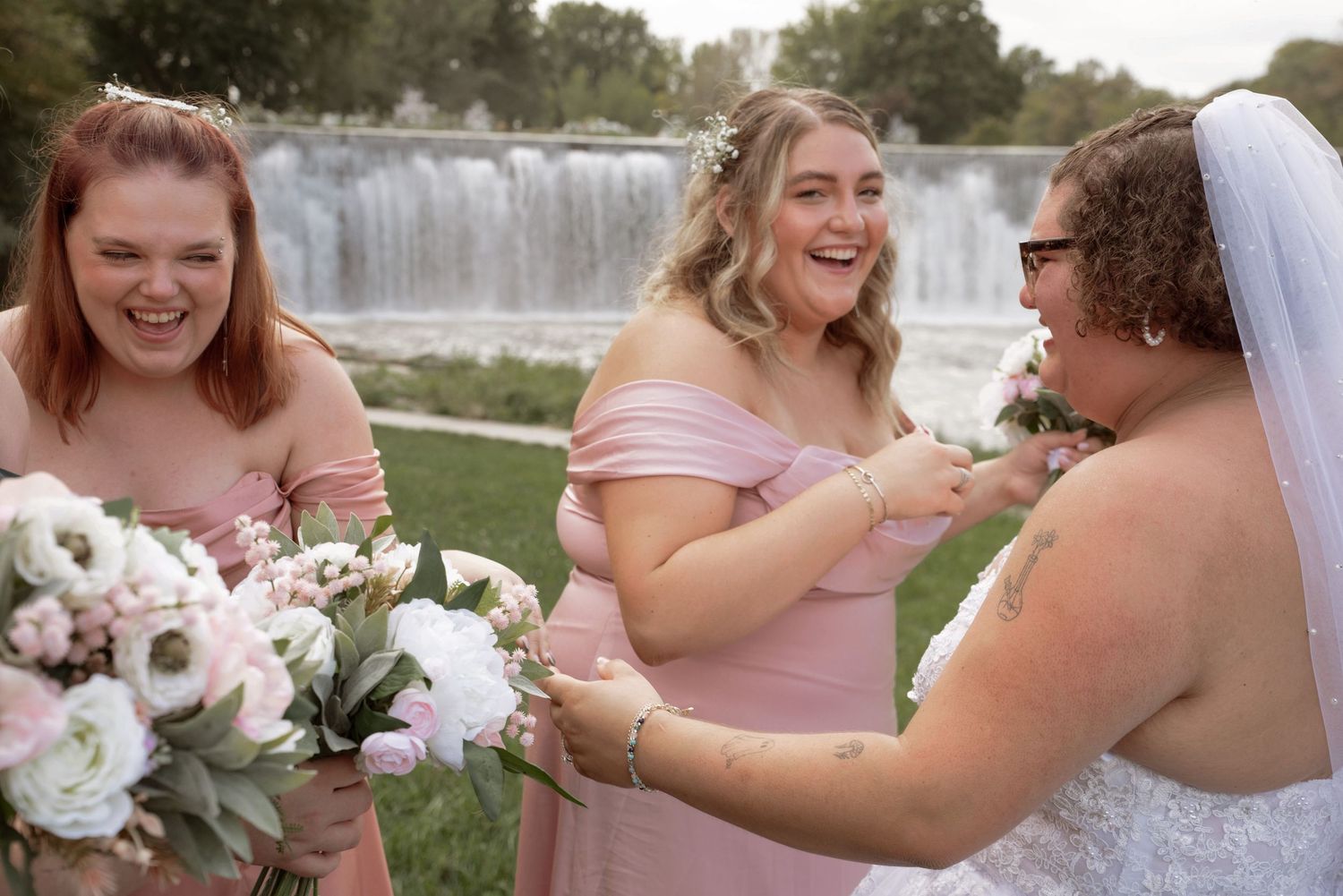 Bridesmaids in pink dresses laughing with bride holding bouquets near a waterfall backdrop in Lanesboro MN.