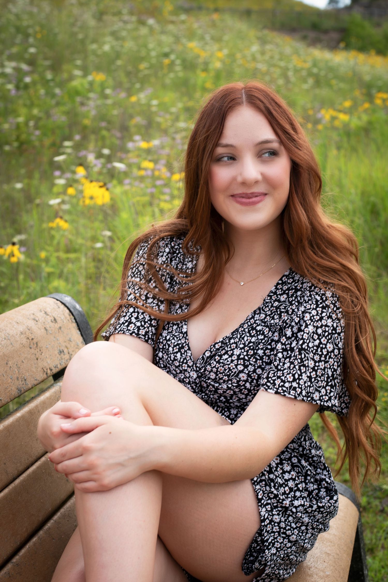 Beautiful redhead teen girl sitting on bench surrounded by colorful wildflowers in floral dress.