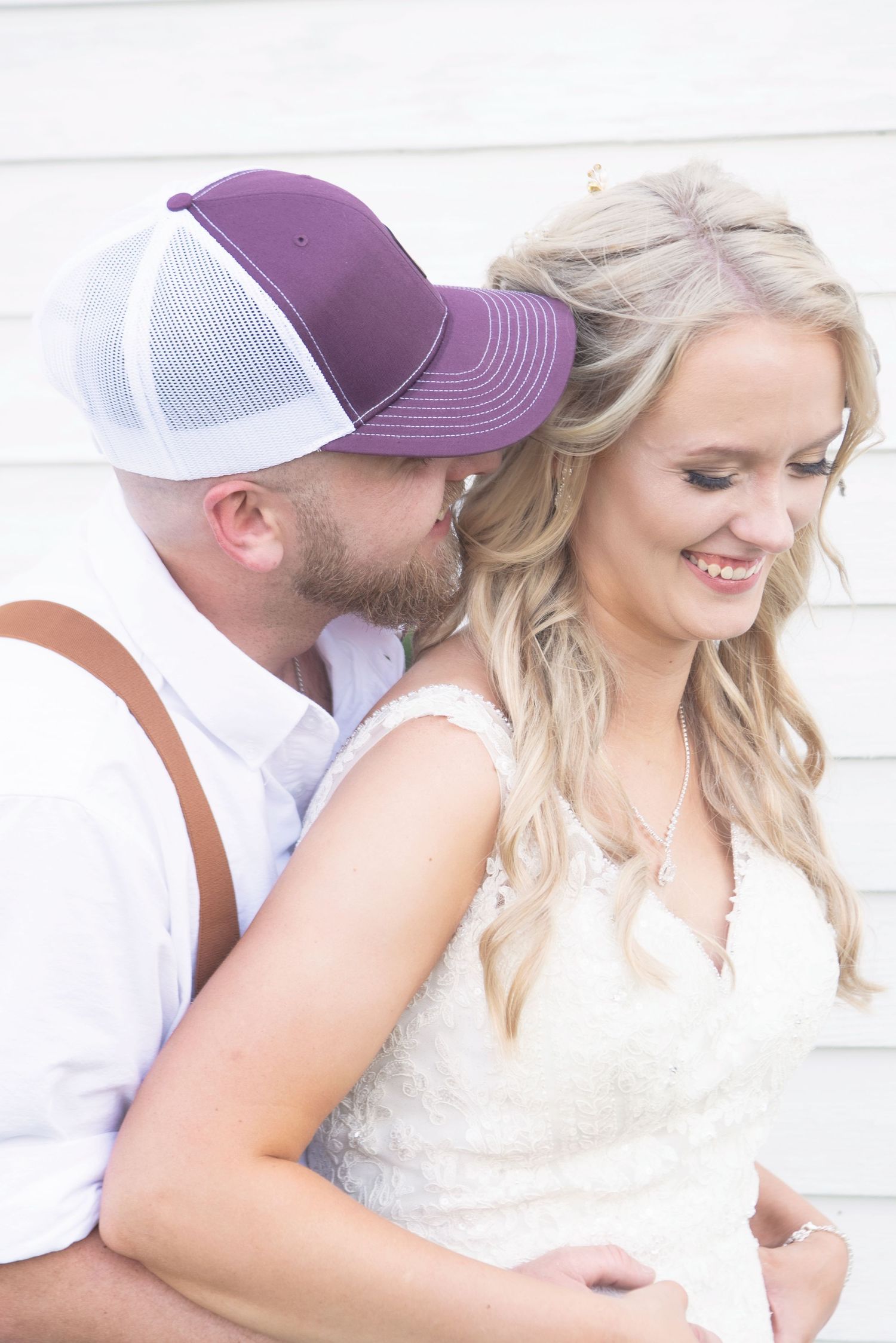 Groom in purple trucker hat kisses smiling bride on the neck against a white wooden backdrop.