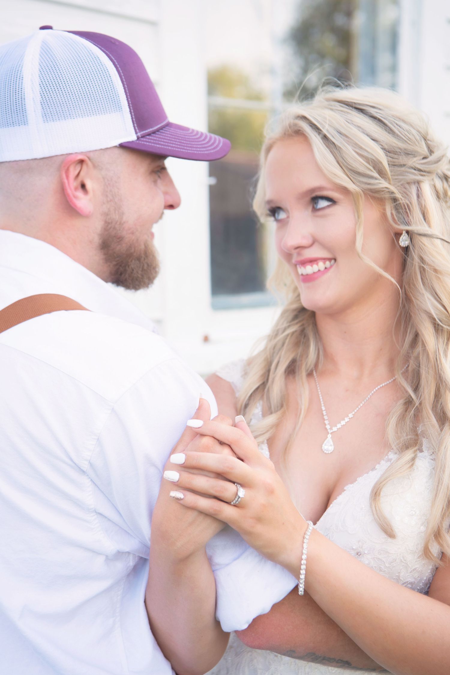 Bride with blonde hair smiles at groom in purple cap and suspenders as they share a tender dance moment.