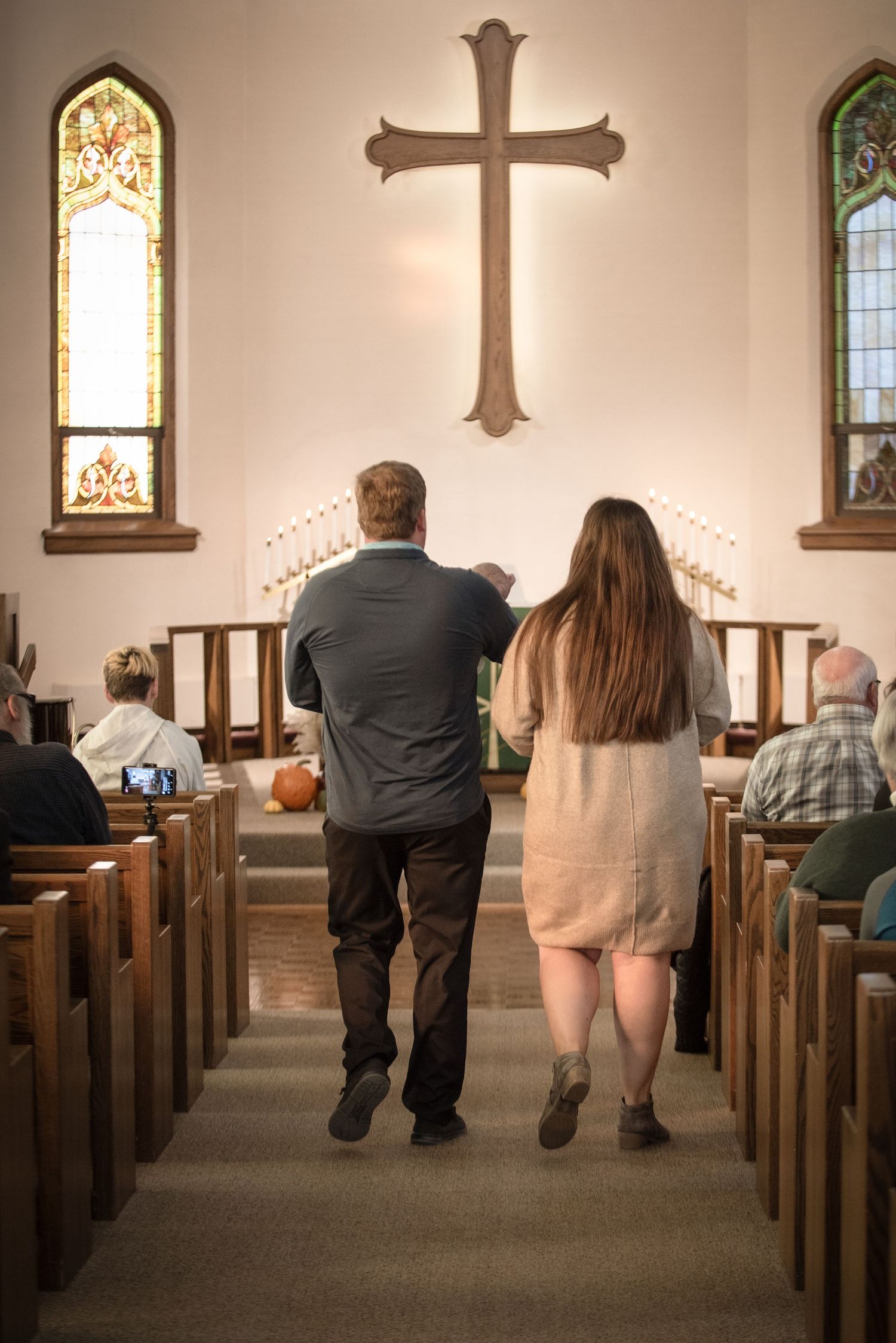 A couple walks hand in hand down the aisle of a traditional church with a wooden cross on the altar.