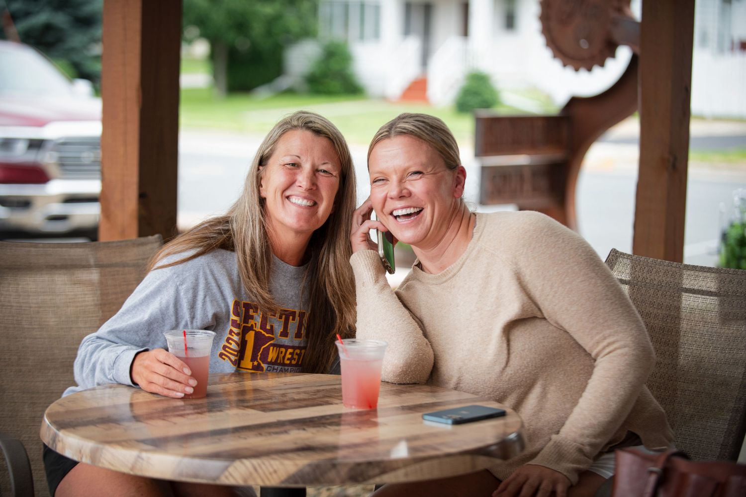 Two smiling women sitting together at a wooden table on an outdoor patio enjoying pink cocktail drinks at Harmony Spirits..