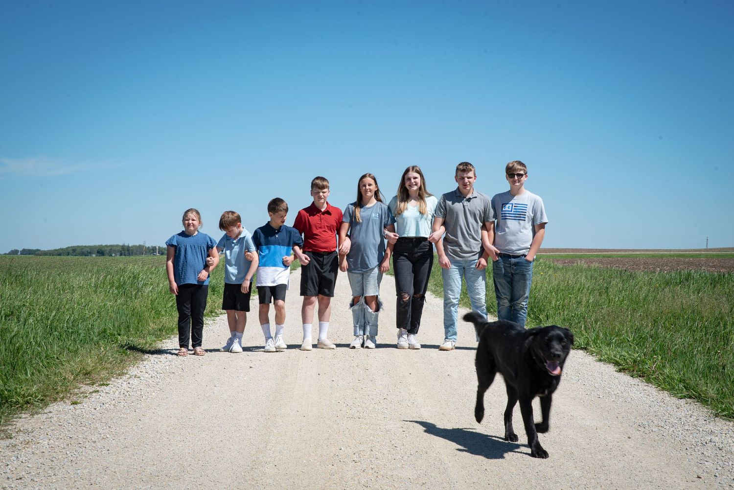 Family with a black dog posing on a gravel farm road under a bright blue sky.