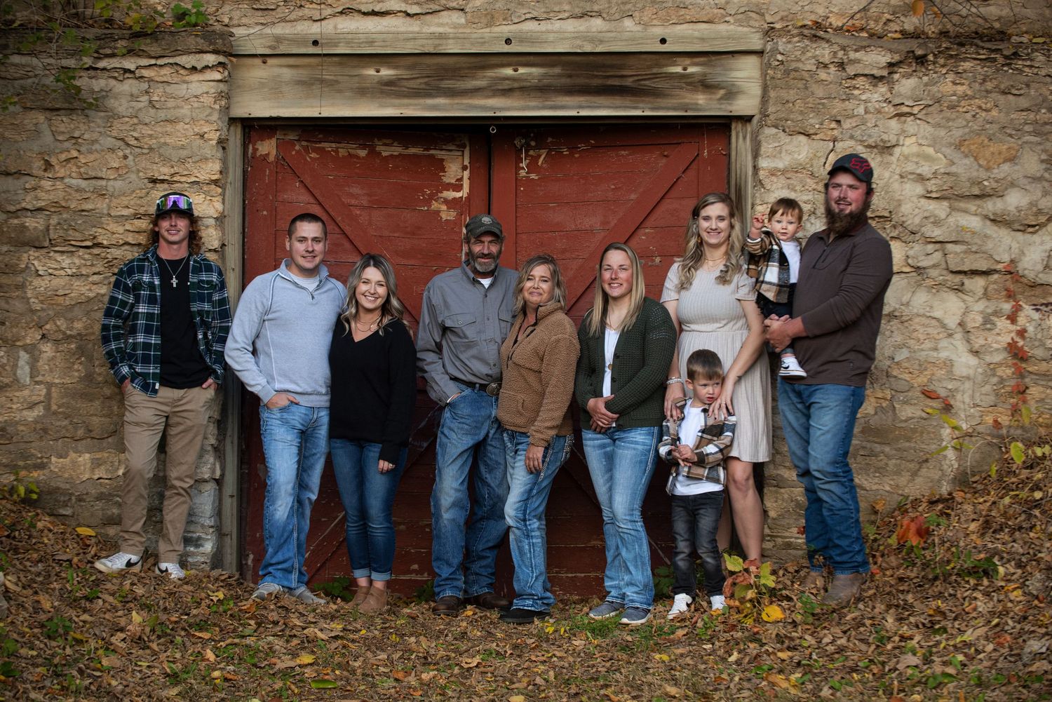 Family of nine poses together in front of a weathered red barn door during autumn outdoor photo session.