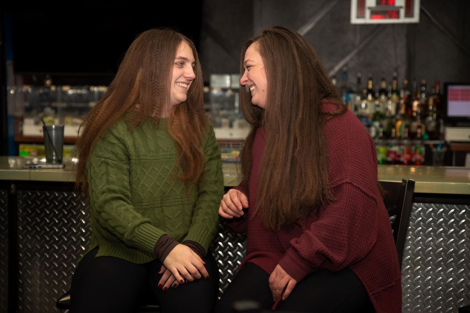 Two young women with long brown hair smile and laugh together while sitting at a bar.