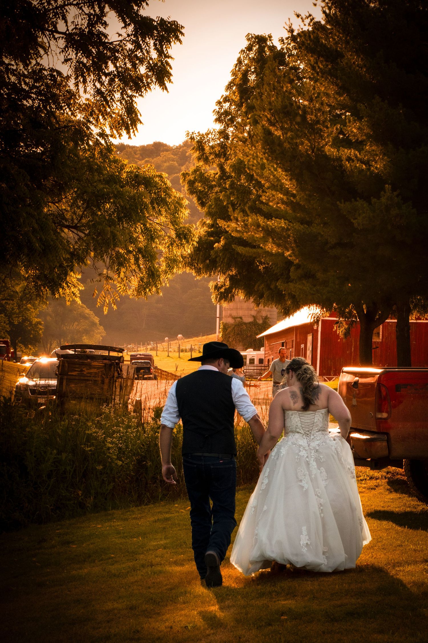 Bride and groom walk hand in hand at golden sunset through rustic farm wedding venue with red barn.