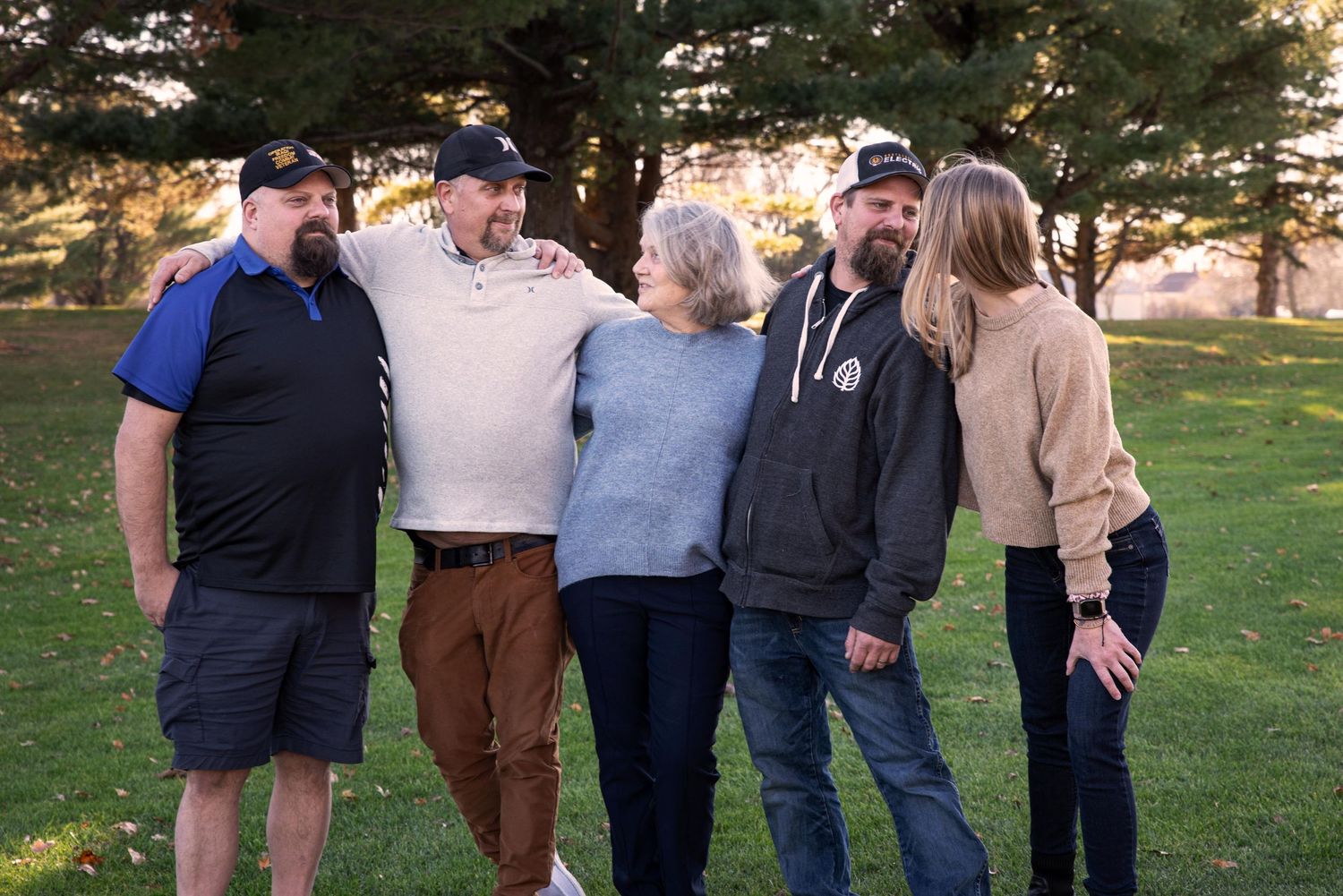 Five family members smile and embrace each other outdoors on a green lawn during a fall family portrait.