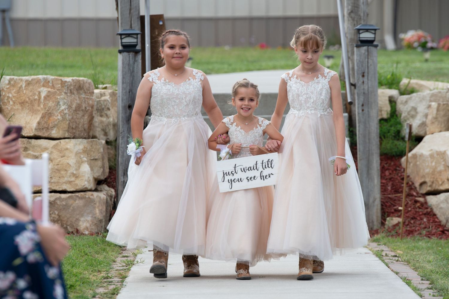 Three flower girls in blush pink tulle dresses walk down outdoor wedding ceremony aisle carrying a decorative sign.