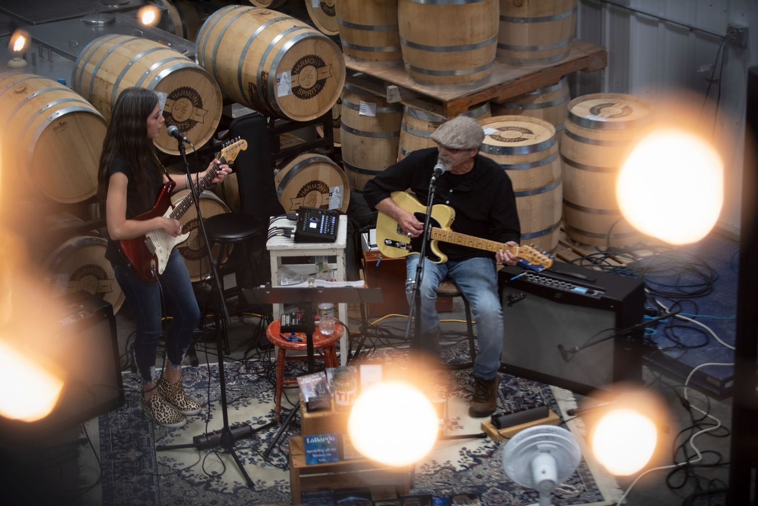 Male and female duo performing live guitar music inside a rustic distillery with oak barrels behind them at Harmony Spirits..