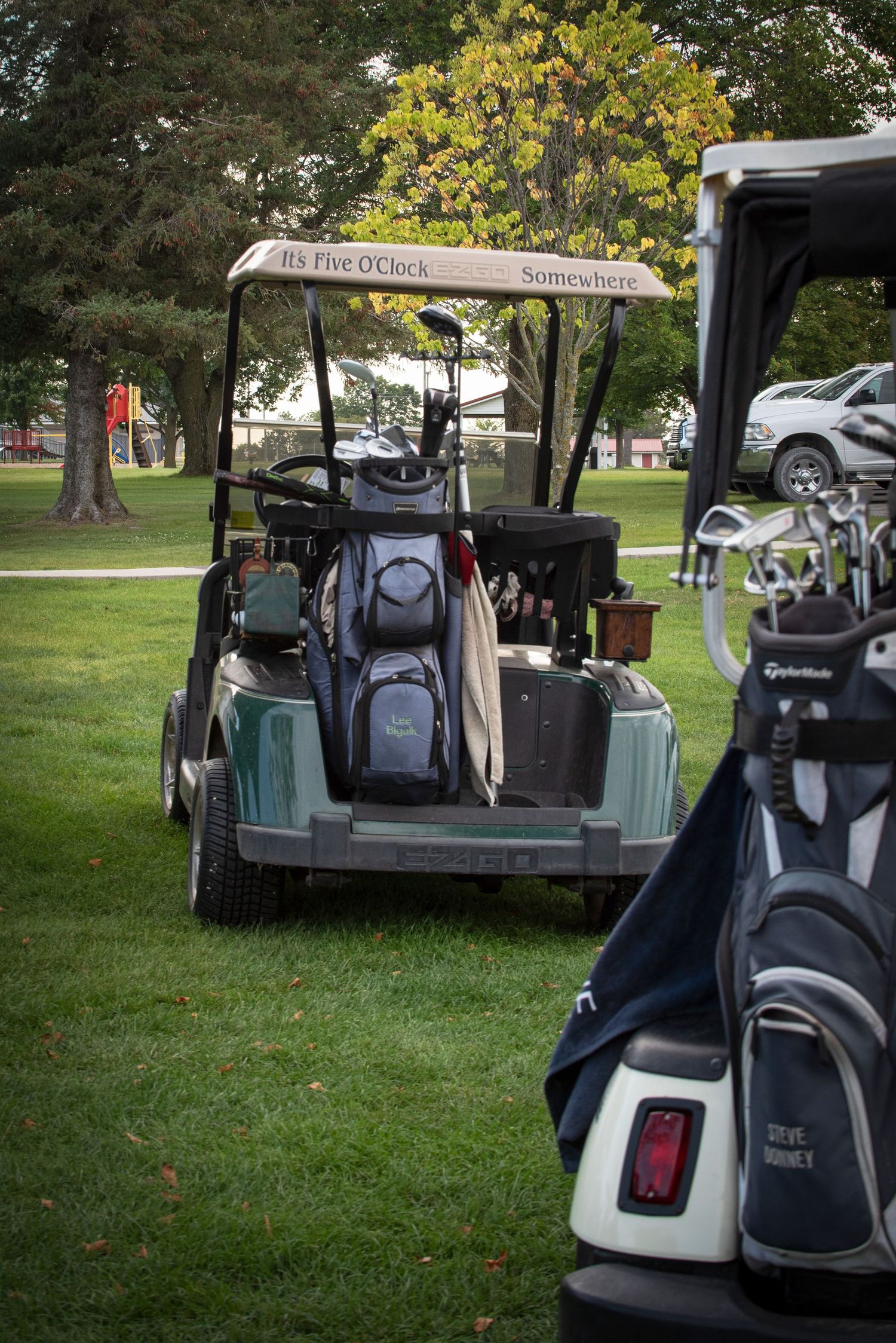 Golf cart with It's Five O'Clock Somewhere sign loaded with golf bags on a green course.