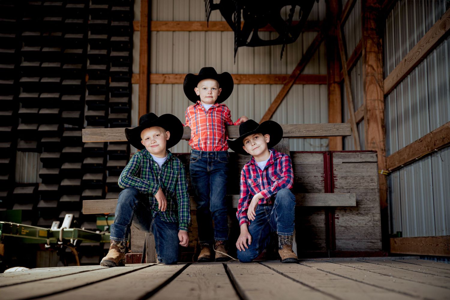 Three young boys wearing black cowboy hats and plaid shirts pose together inside a rustic barn.