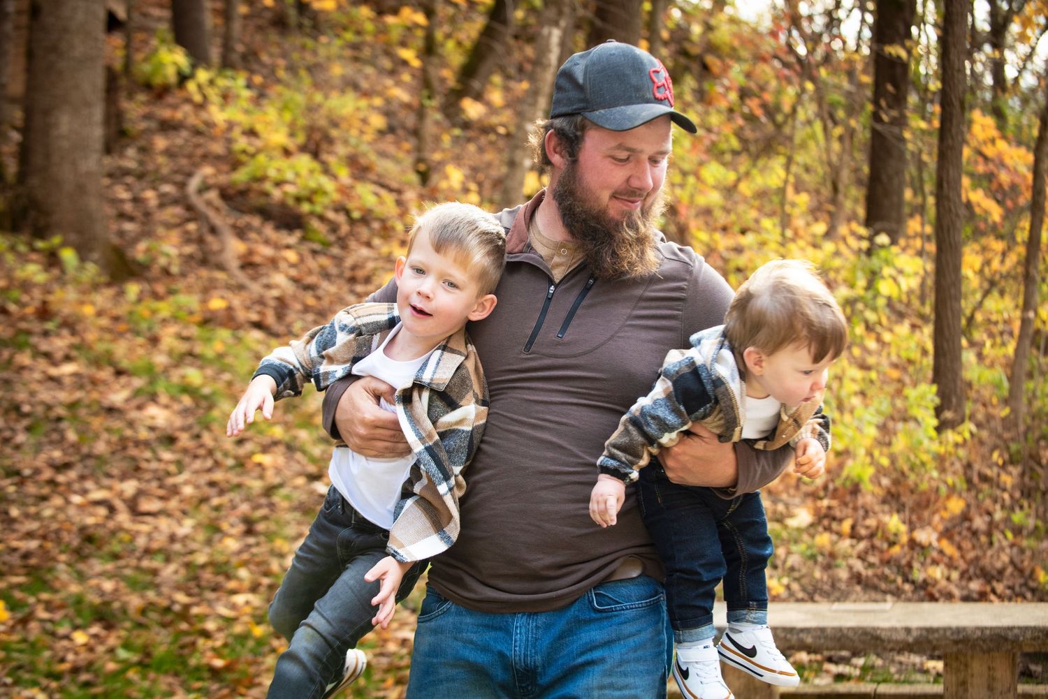 Bearded father carries two laughing young boys on his arms along a wooded autumn trail with fall leaves.