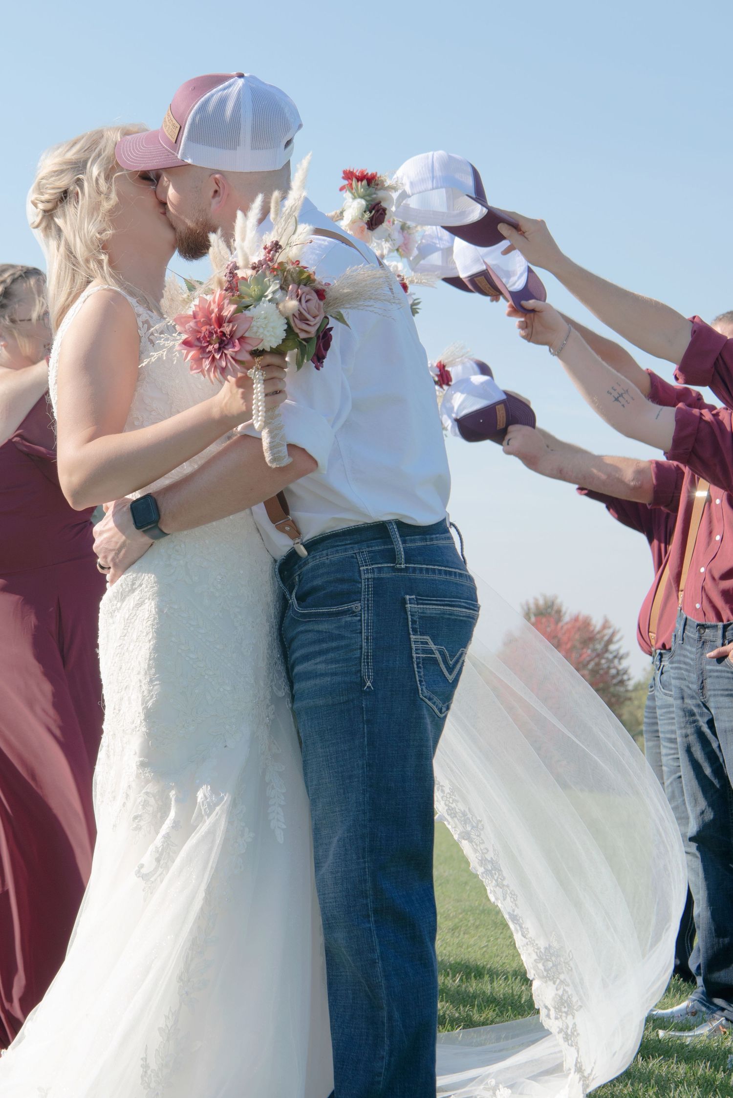 Bride and groom share a kiss under an arch of raised hats formed by wedding party in matching maroon caps.
