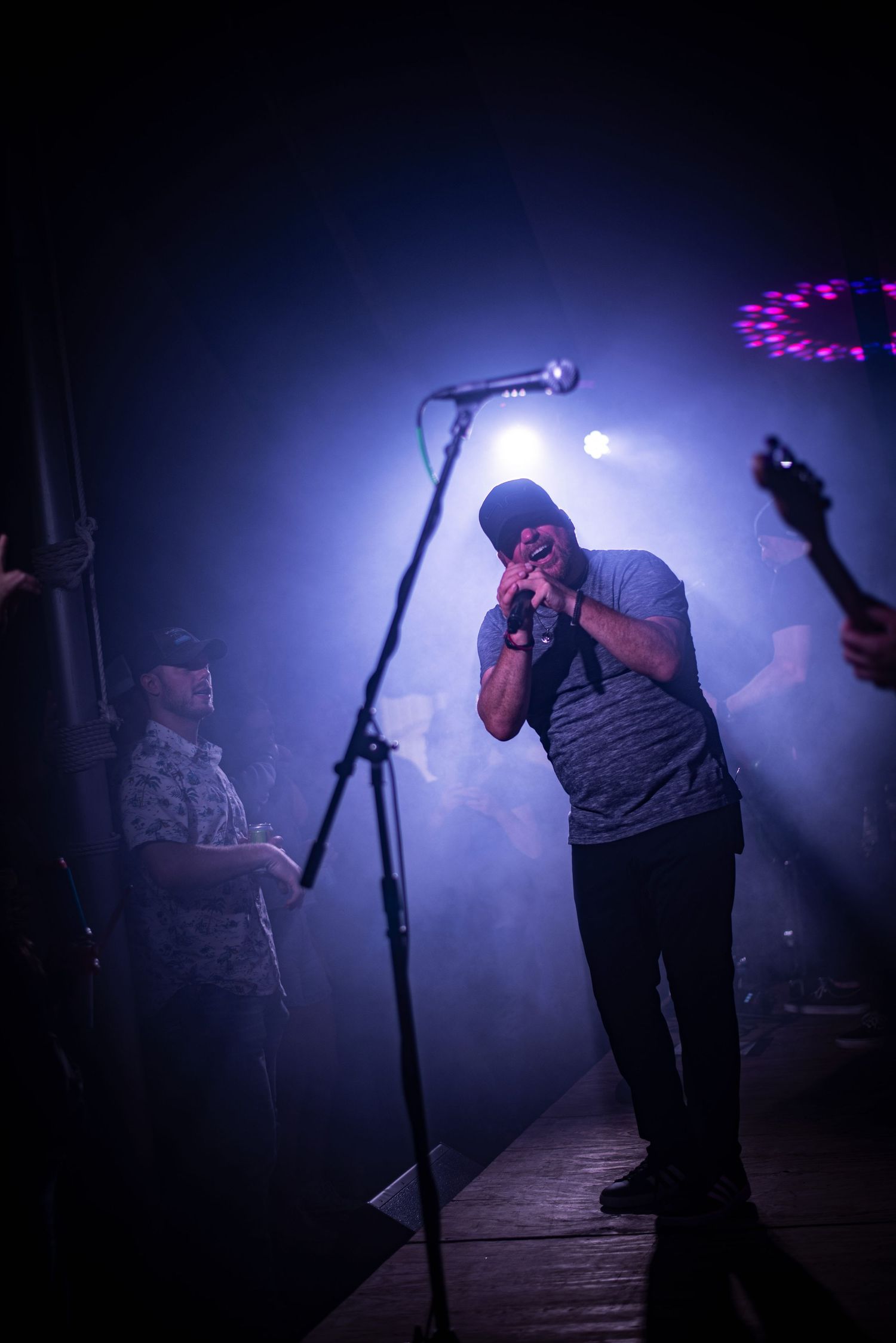 Male vocalist performing on a foggy stage under blue and purple lights with band members beside him.