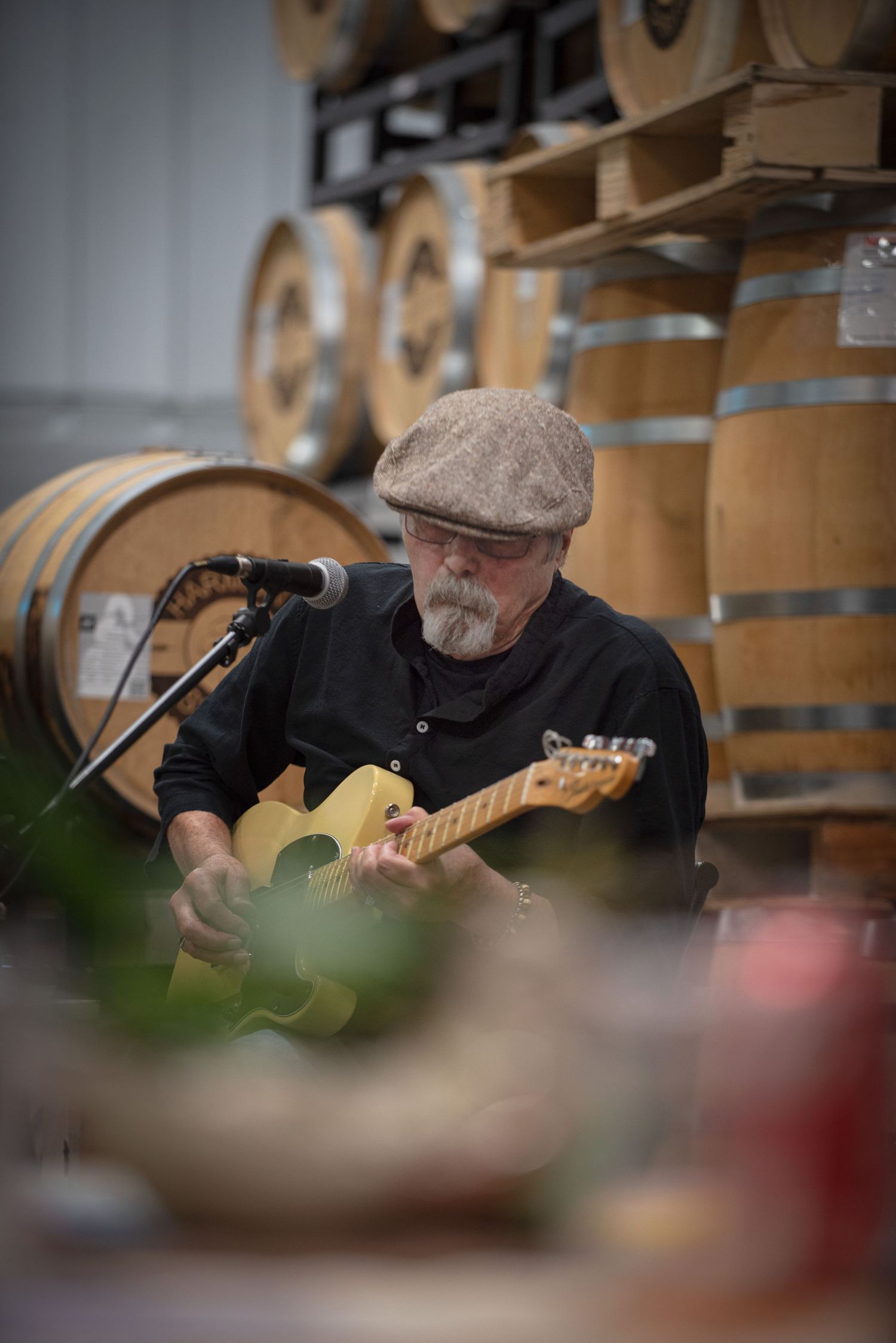 Elderly man playing yellow electric guitar at a winery surrounded by oak barrels at Harmony Spirits..