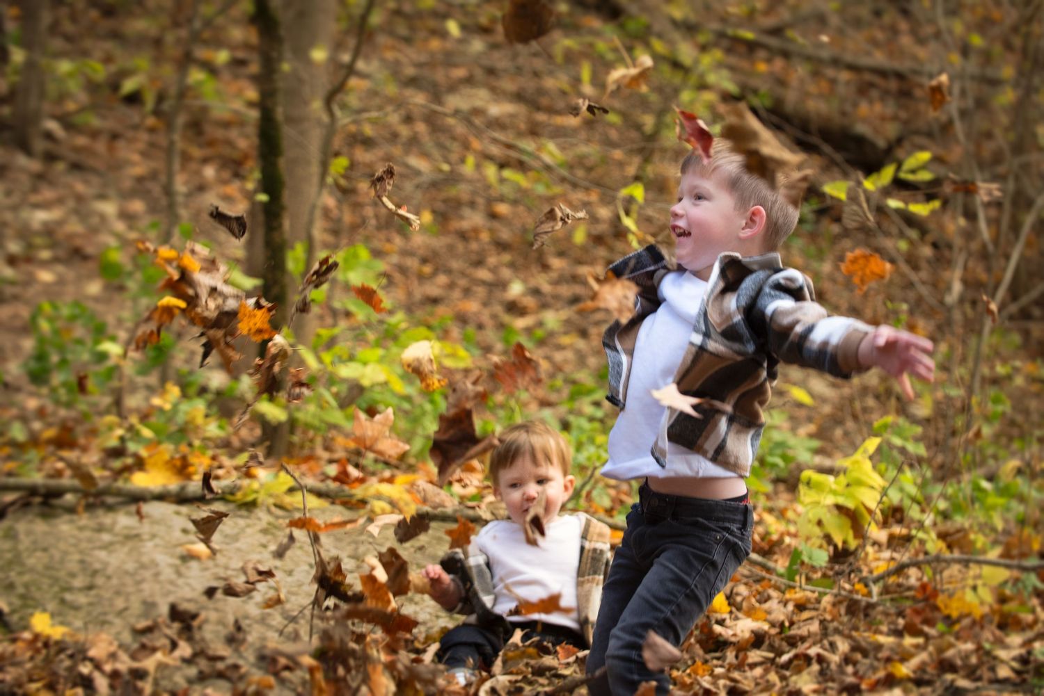 Two young boys joyfully tossing autumn leaves in a woodland setting.