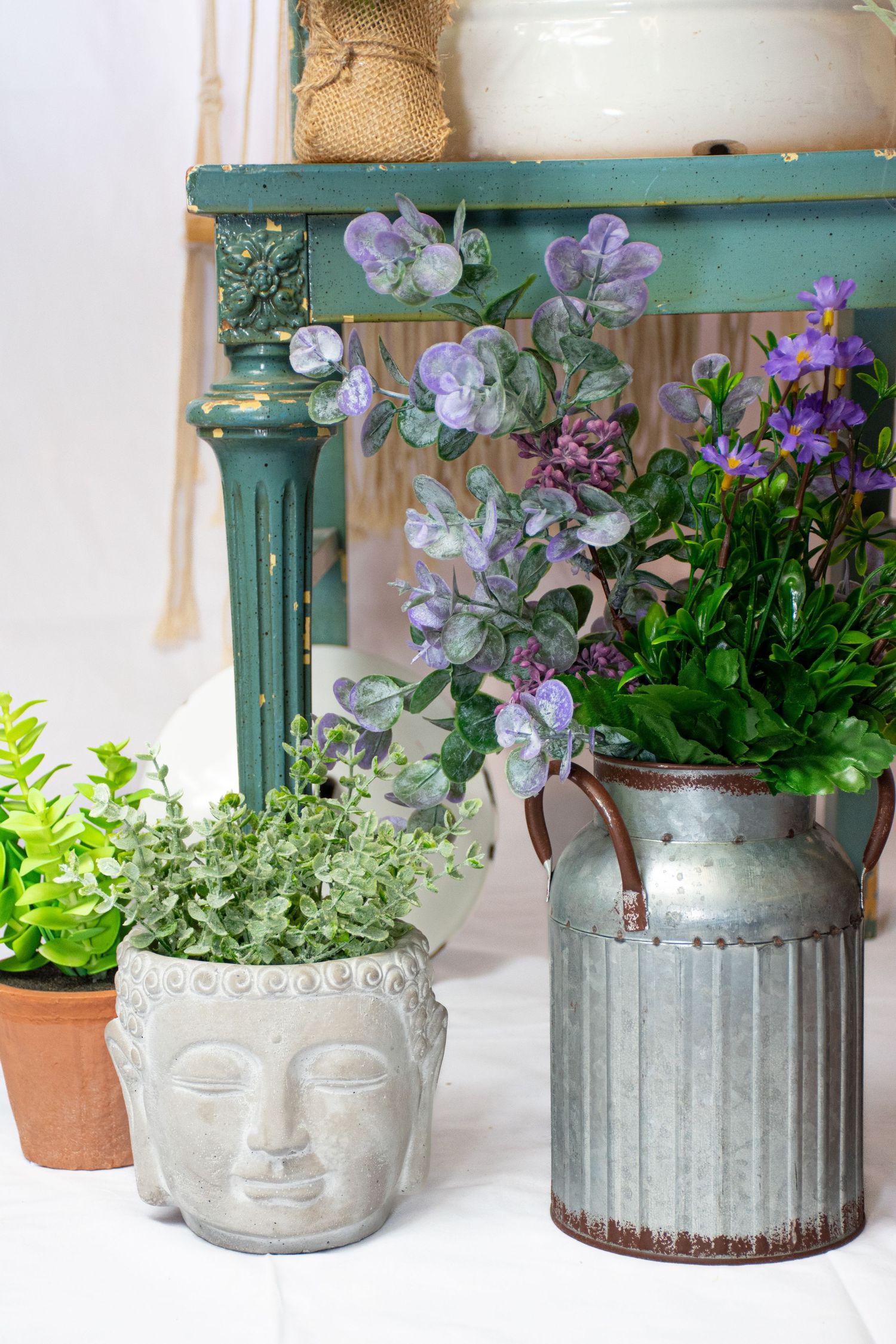 Purple flowers and green plants in Buddha head pot and rustic metal milk can near teal table at Bee Balm Boutique.
