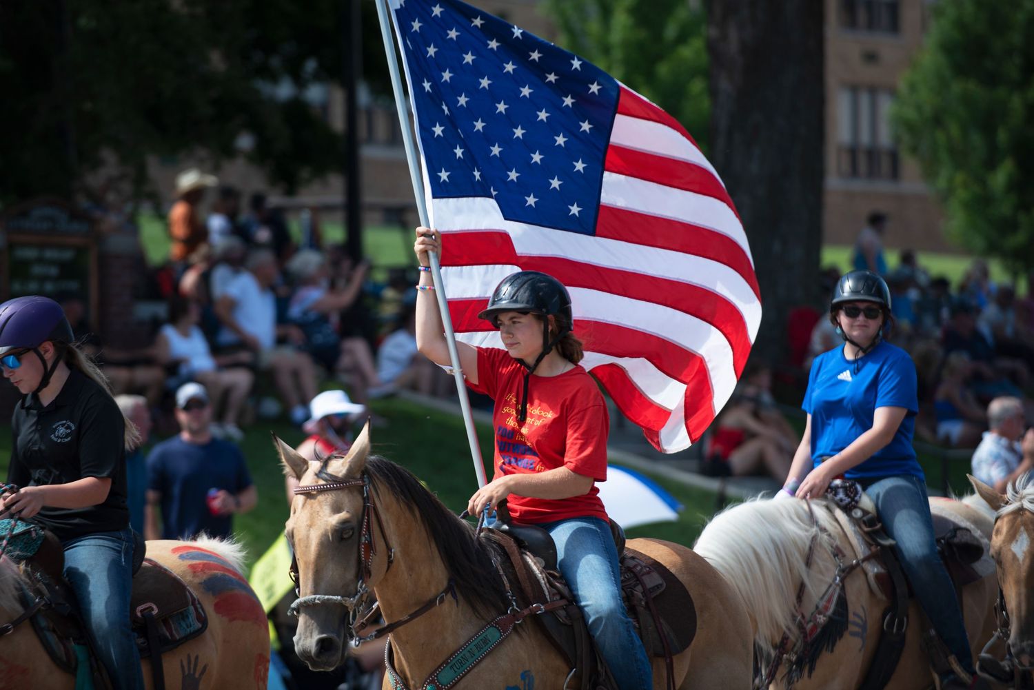 Person on horseback carrying large American flag in outdoor parade with crowd watching on sunny day at Harmony MN 4th of July Celebration..