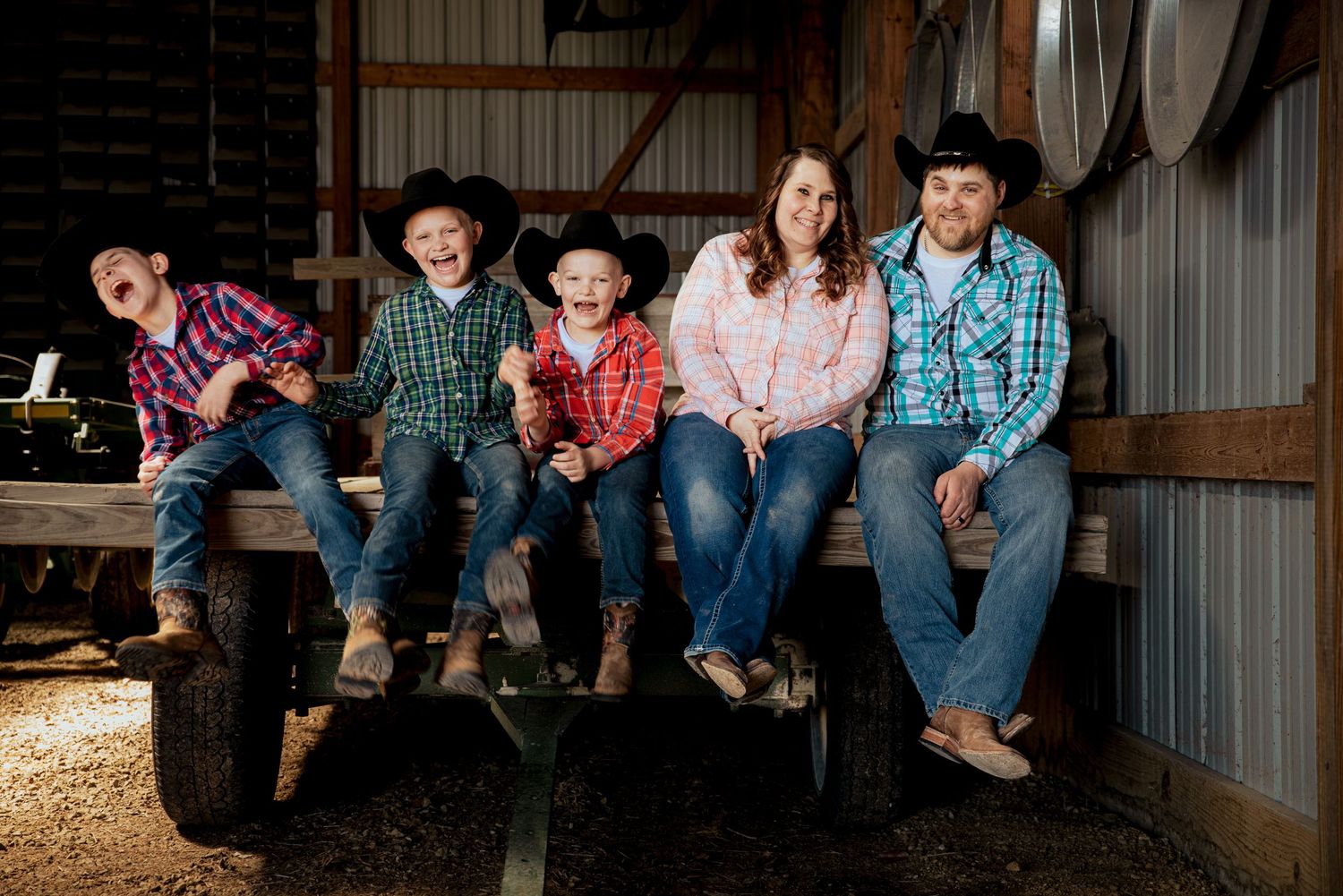 Family of five in plaid shirts and cowboy hats sitting together laughing on a wooden farm wagon.