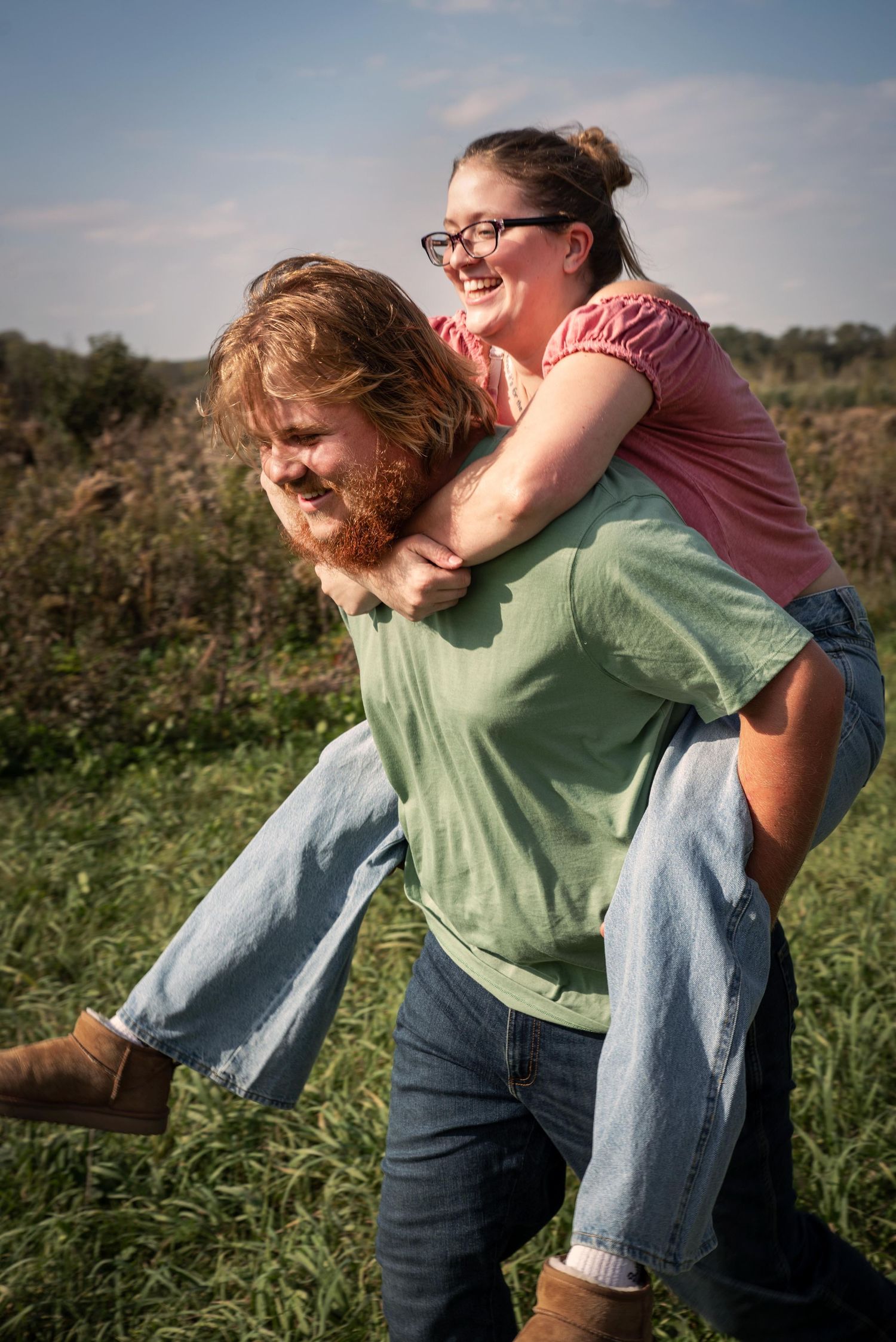 Joyful couple laughing as man gives woman a piggyback ride through a sunny meadow.