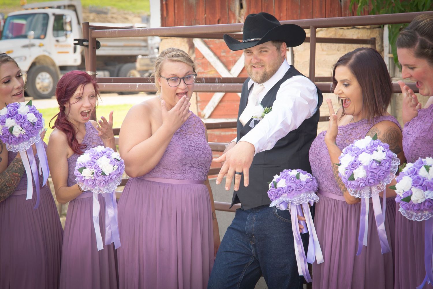 Groom in black cowboy hat playfully poses with surprised bridesmaids in lavender dresses at rustic barn wedding.