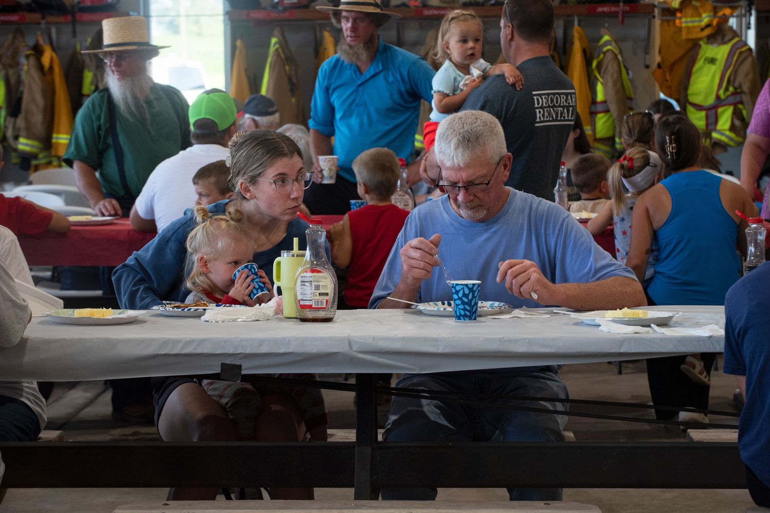 Families gather at picnic tables inside a fire station for a community meal event. 4th of July Celebration in Harmony MN.
