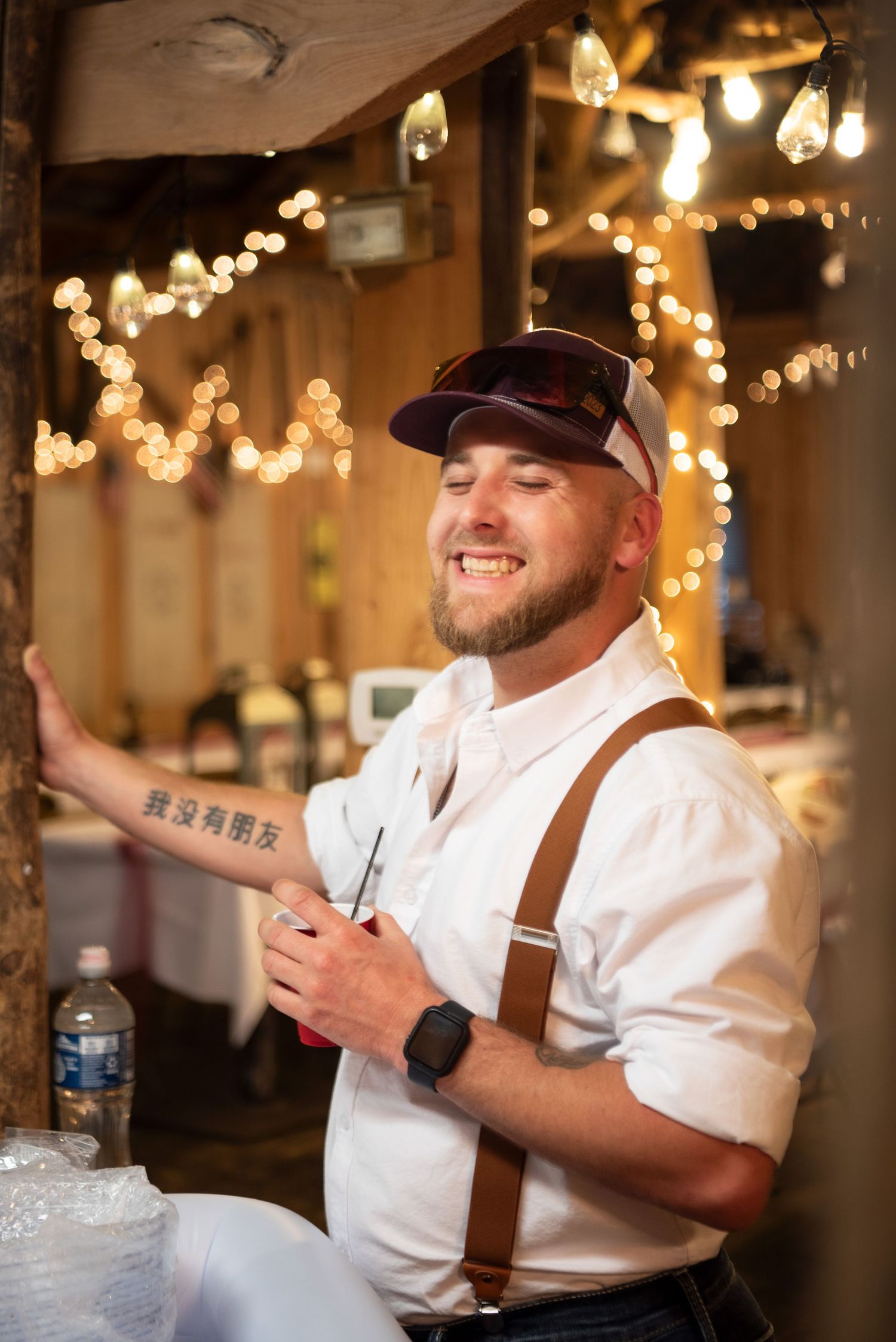 Smiling groom in white shirt, suspenders, and baseball cap holds pen at rustic barn wedding reception with string lights.