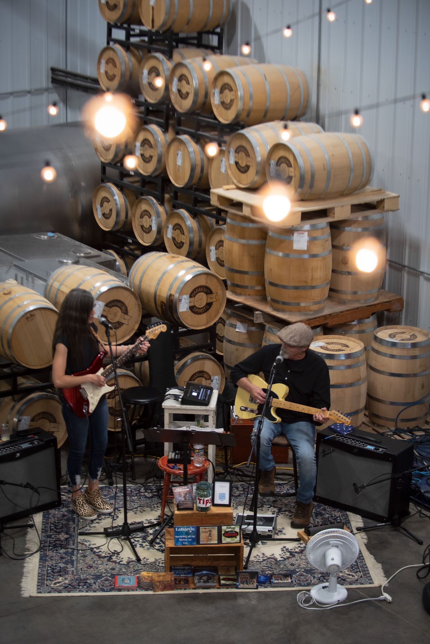 Two musicians performing live guitar music inside a distillery surrounded by stacked oak aging barrelsat Harmony Spirits..