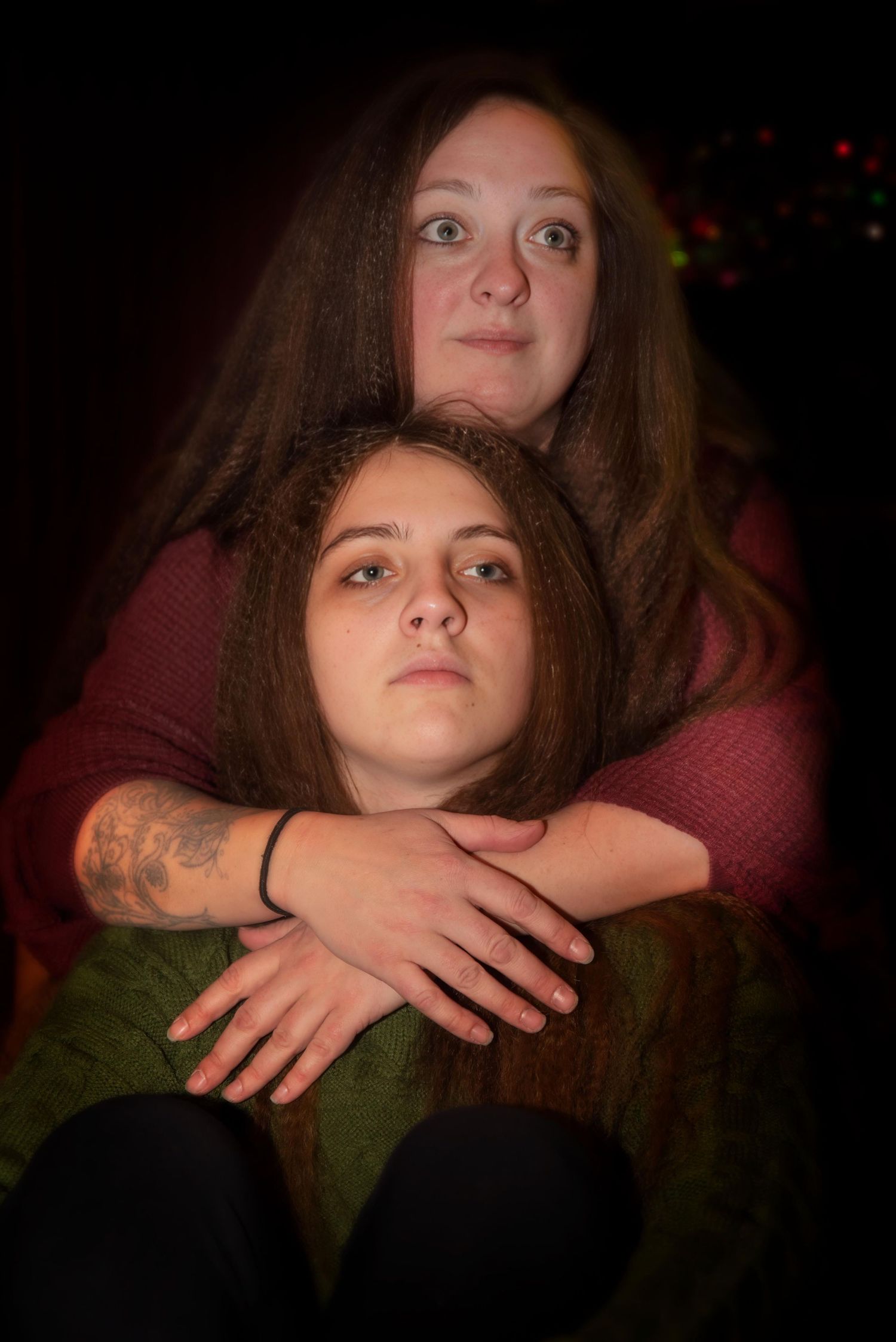 Two brown-haired women in emotional embrace pose against dark background with bokeh lights.