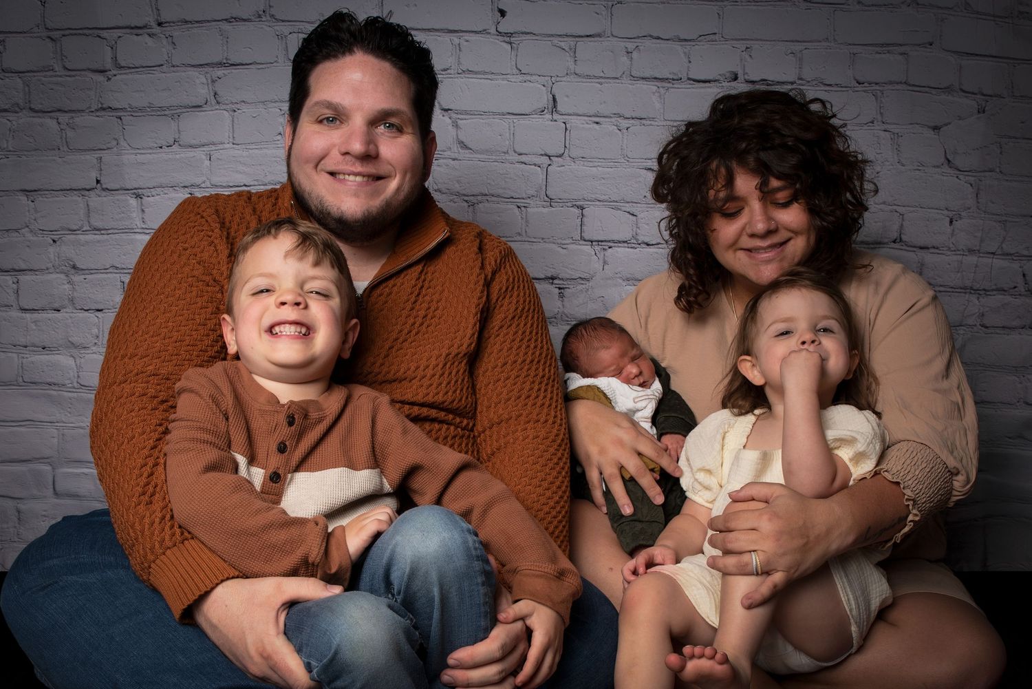 Happy family of five with parents holding three children including a newborn posed against a brick wall.
