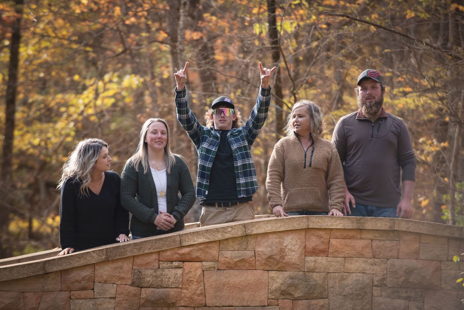 Family of five stands on a stone bridge wall with one member pointing upward among autumn colored trees.